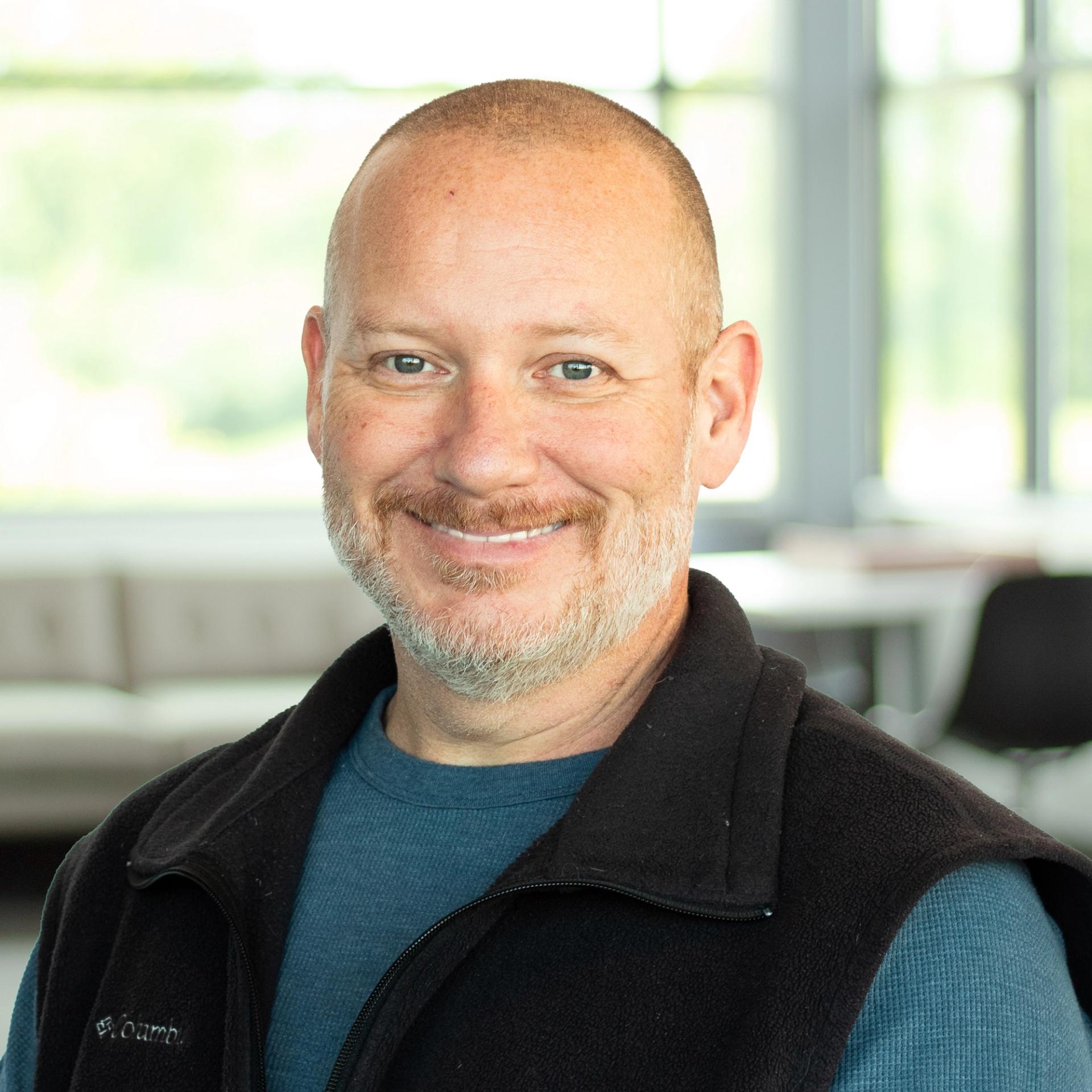 Garry Graves, wearing a blue long-sleeve shirt and black Columbia vest, smiling in a bright, modern office.