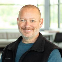 Garry Graves, wearing a blue long-sleeve shirt and black Columbia vest, smiling in a bright, modern office.