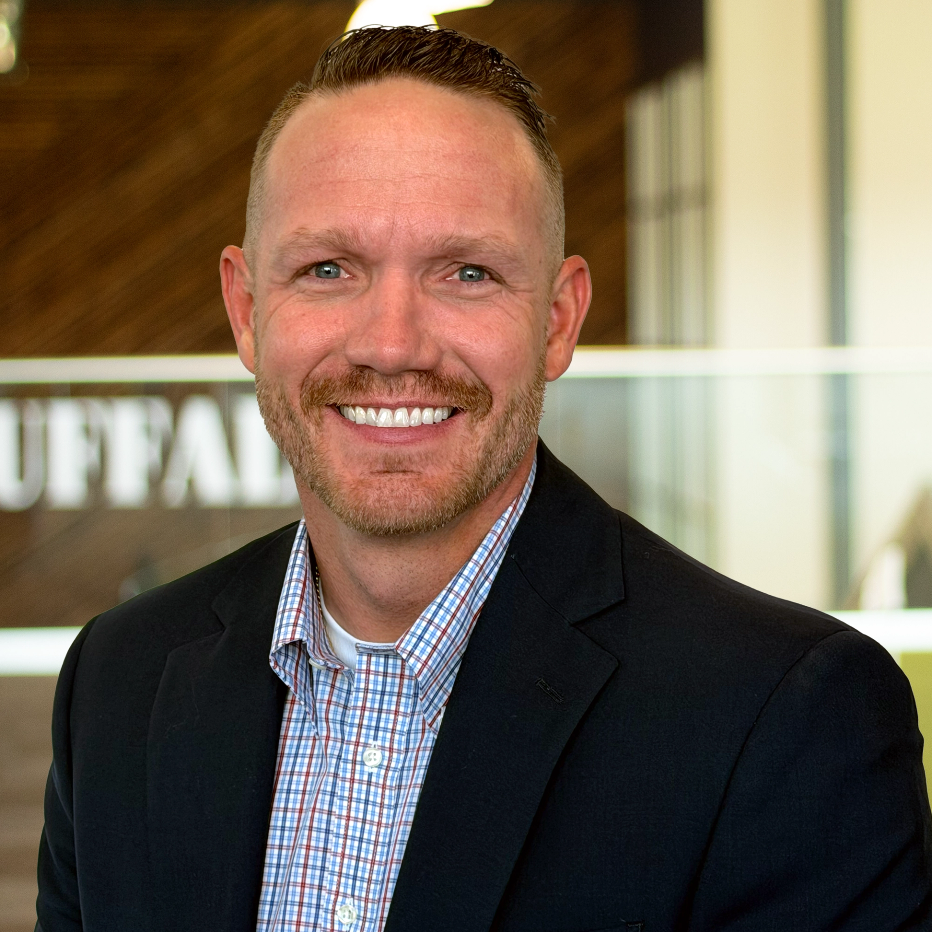 Stephen Herzog wearing a checked dress shirt and navy blazer, standing in front of a company-branded wall.