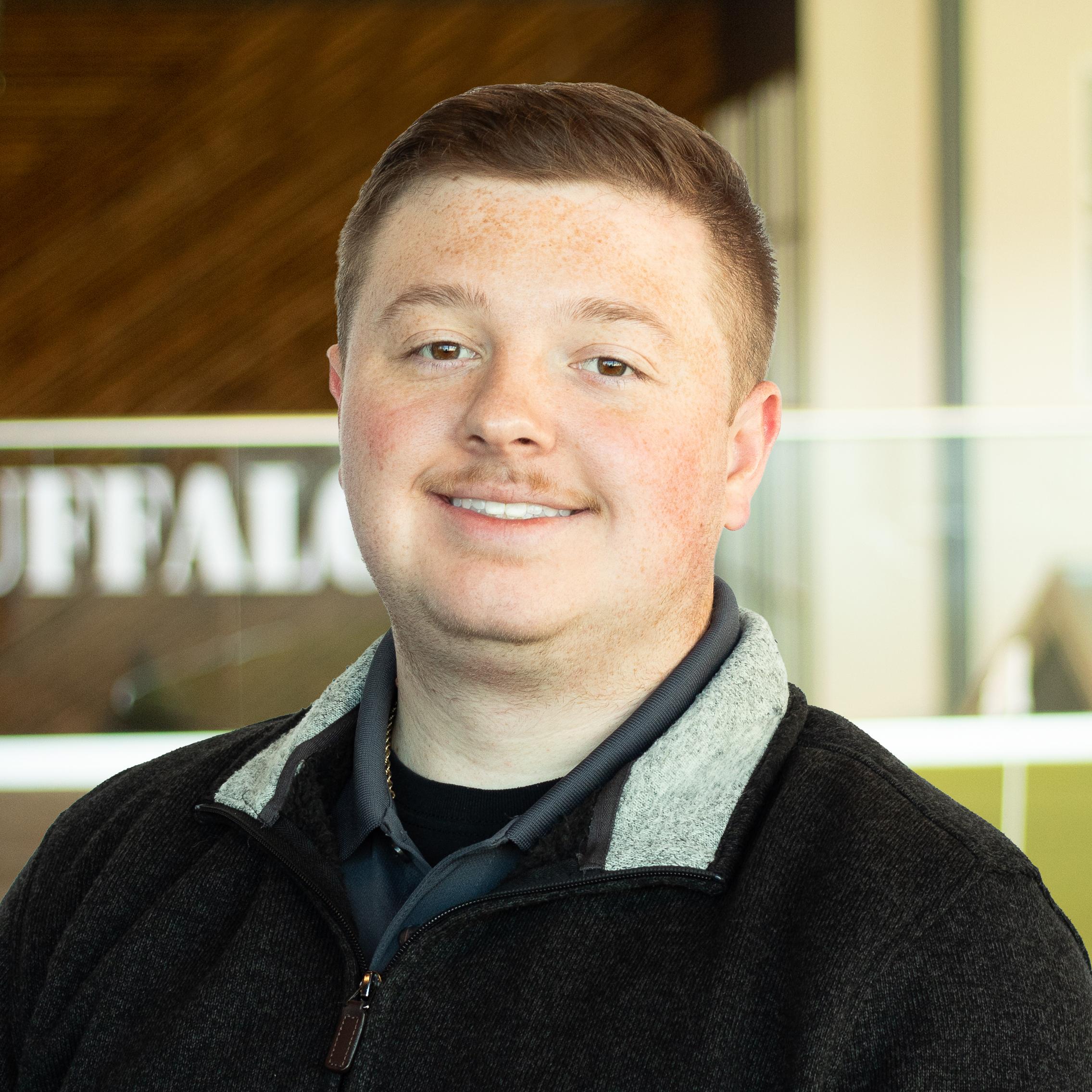 Charlie Fowler, smiling, wearing a dark zip-up fleece with a collared shirt underneath, standing in Buffalo’s office space.