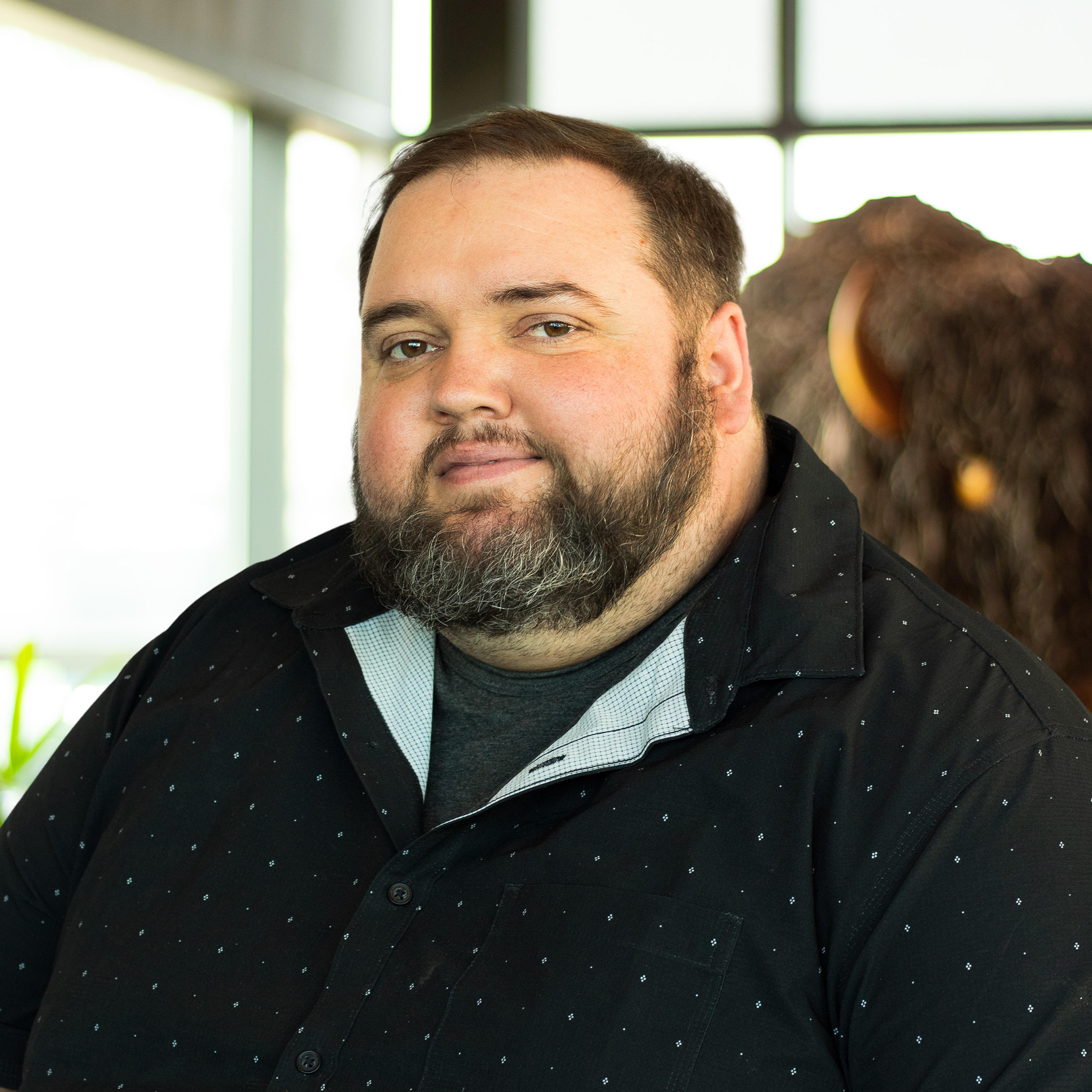 Justin Rhodes smiles while standing in front of a buffalo statue inside the Buffalo Construction office, wearing a light gray zip-up jacket over a black shirt.