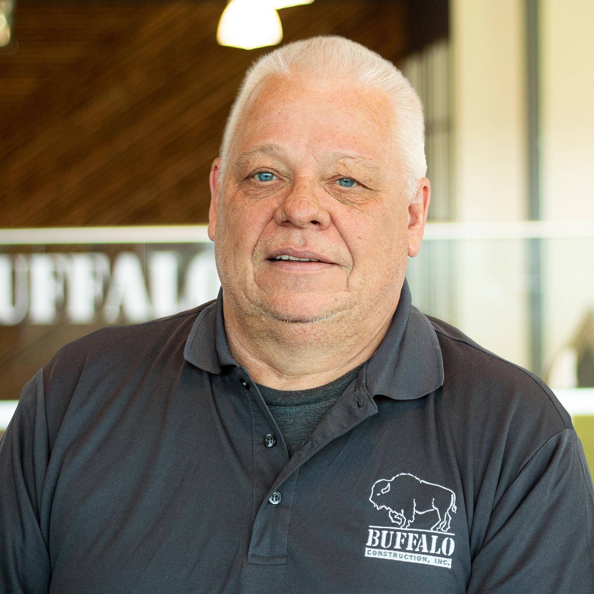 Larry Glidewell with a faint smile, wearing a buffalo construction branded polo and standing in buffalo's office space.