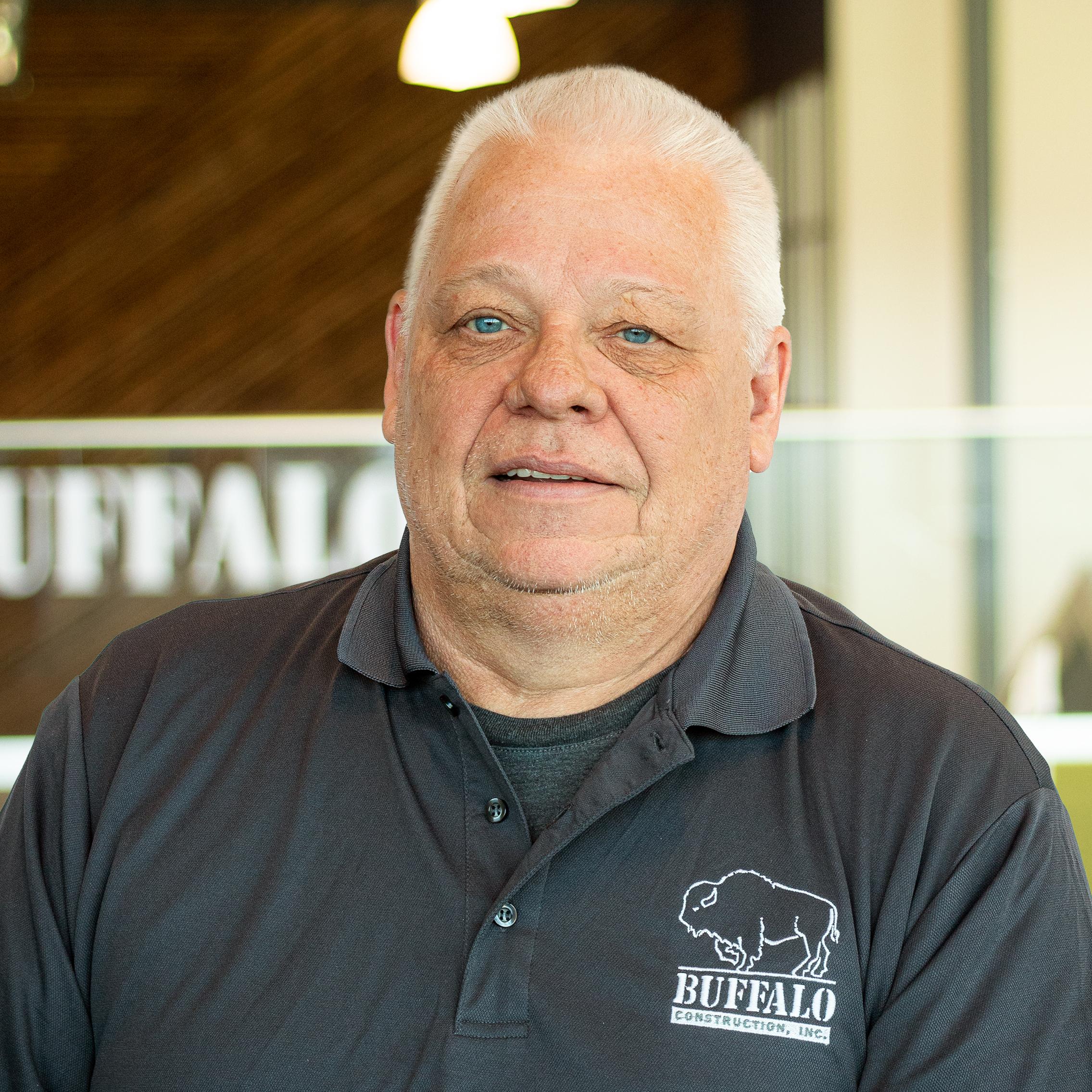 Larry Glidewell with a faint smile, wearing a buffalo construction branded polo and standing in buffalo's office space.