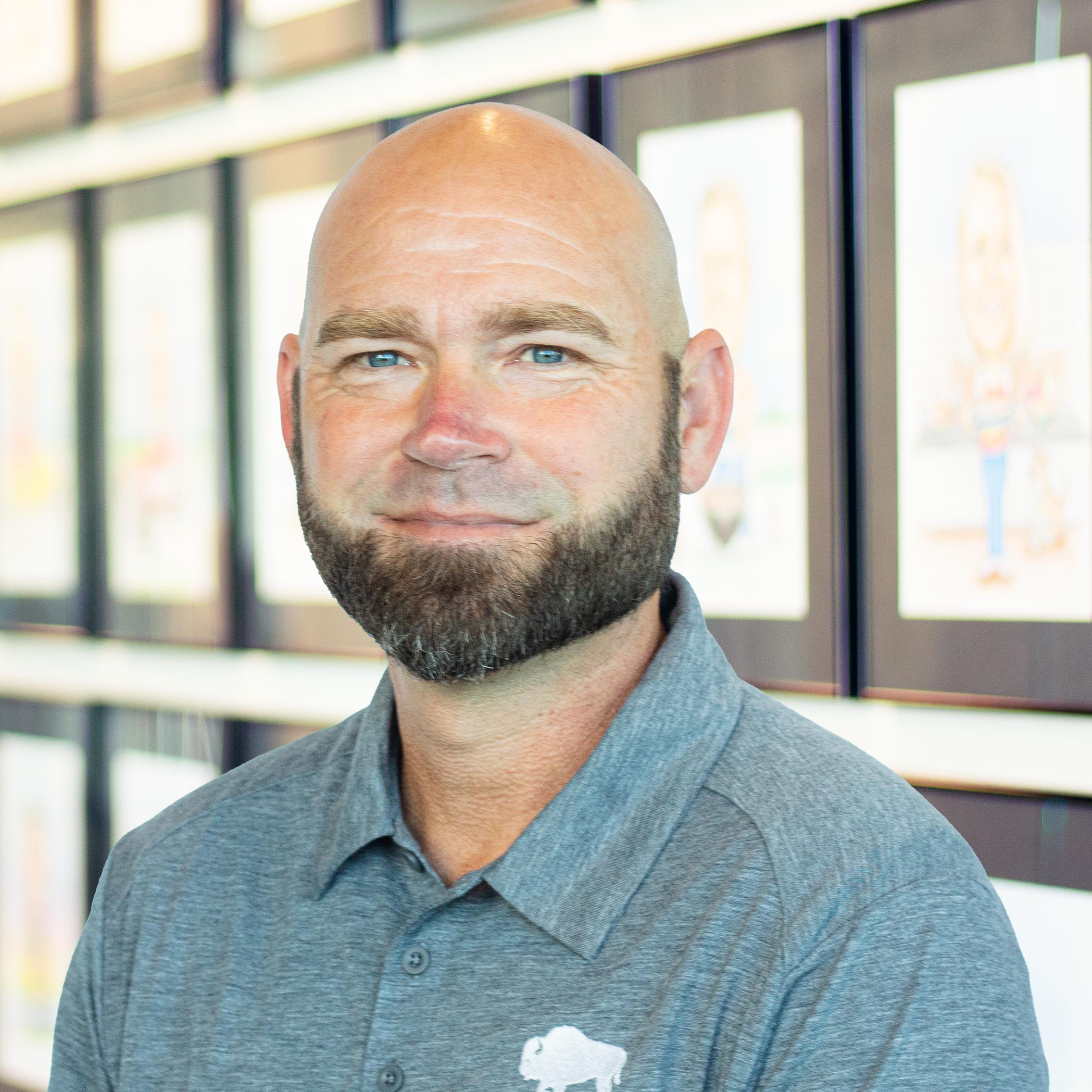 Bald man with a full beard wearing a gray polo with a buffalo logo, standing before a wall of framed caricatures.