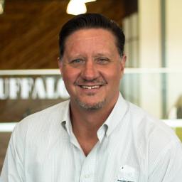 Robert Burns wearing a light button-down shirt and standing in front of a company-branded wall.