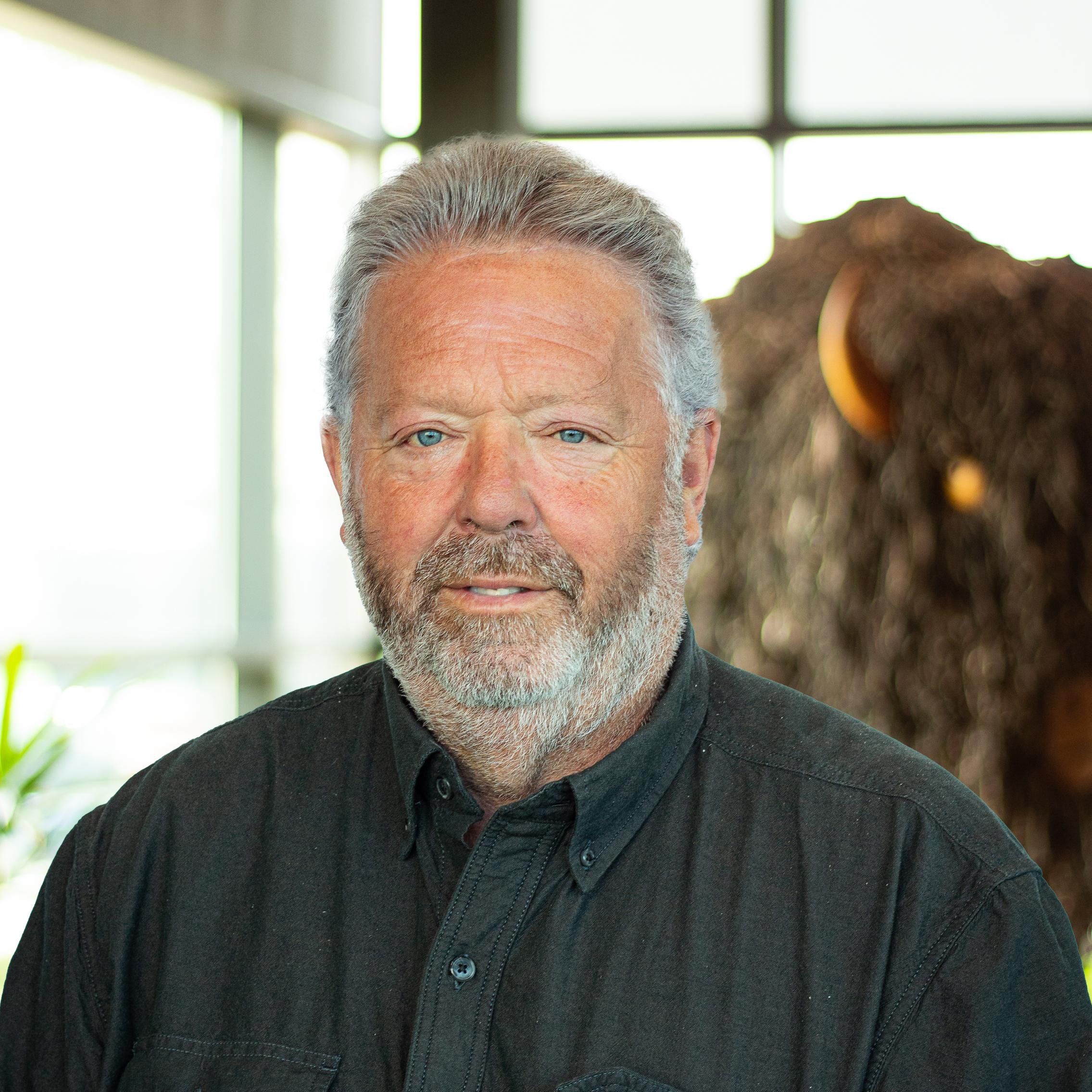 Older man with a gray beard and light hair, wearing a dark shirt, standing in front of a buffalo sculpture.