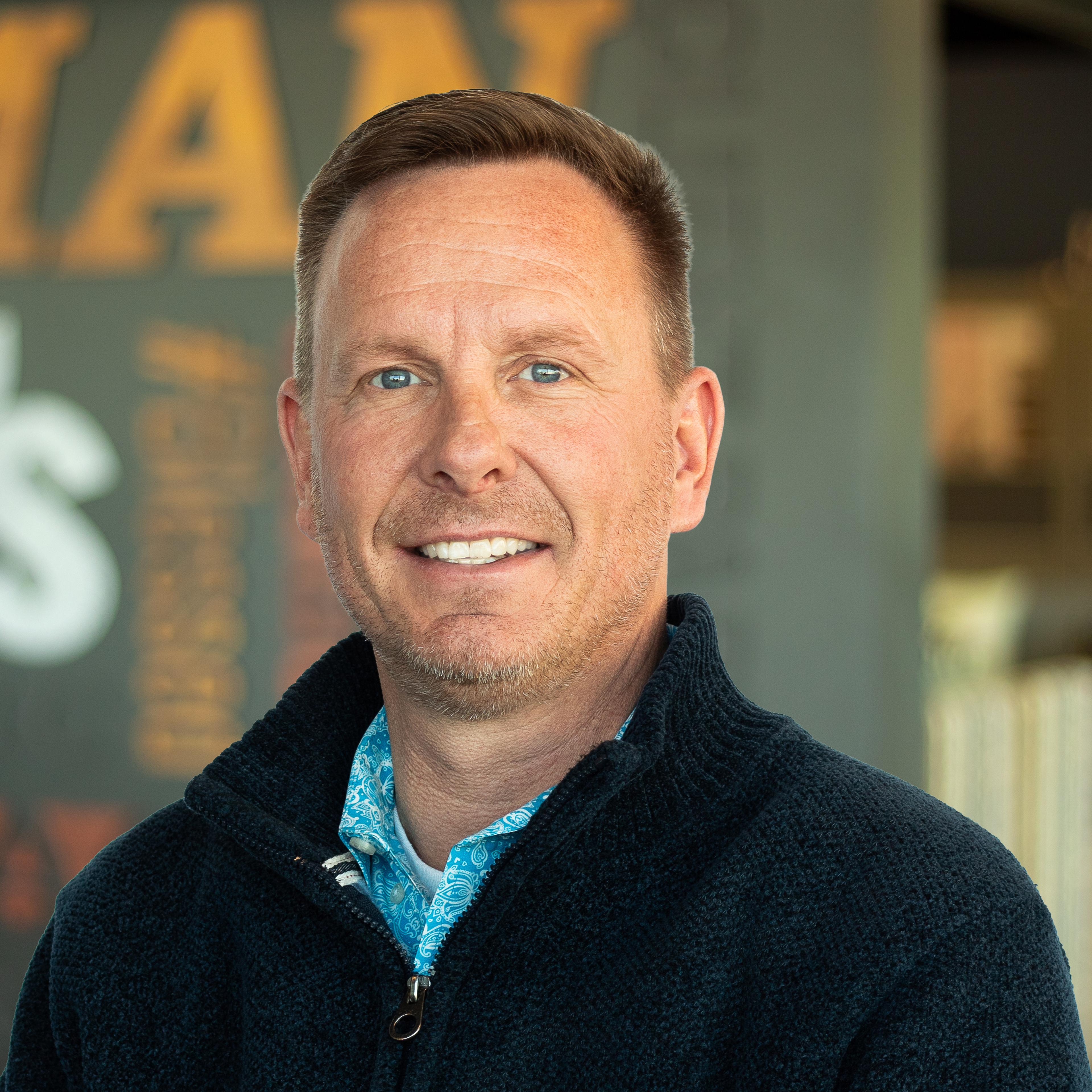 Jason Crow, wearing a textured navy zip-up and blue paisley button-down shirt, standing in front of a company-branded wall.
