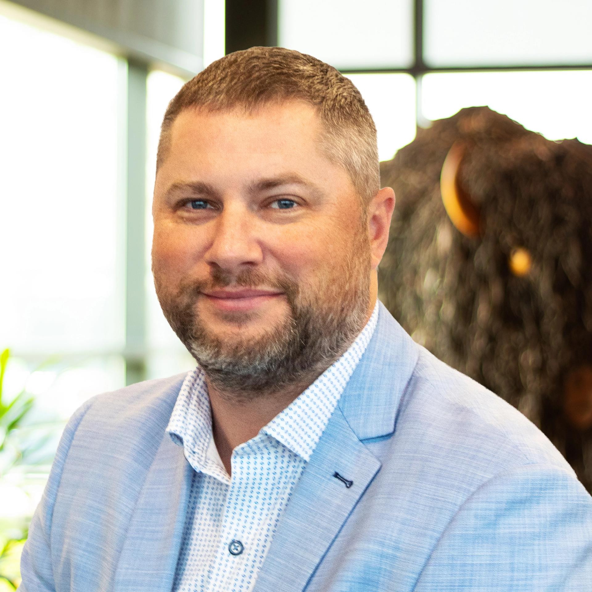 Tim Fishburn smiling, wearing a light blue blazer, with a statue of a buffalo behind him.