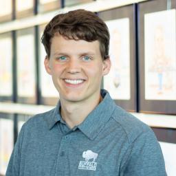 Young man with short brown hair and blue eyes in a gray Buffalo polo, smiling in front of framed illustrations.