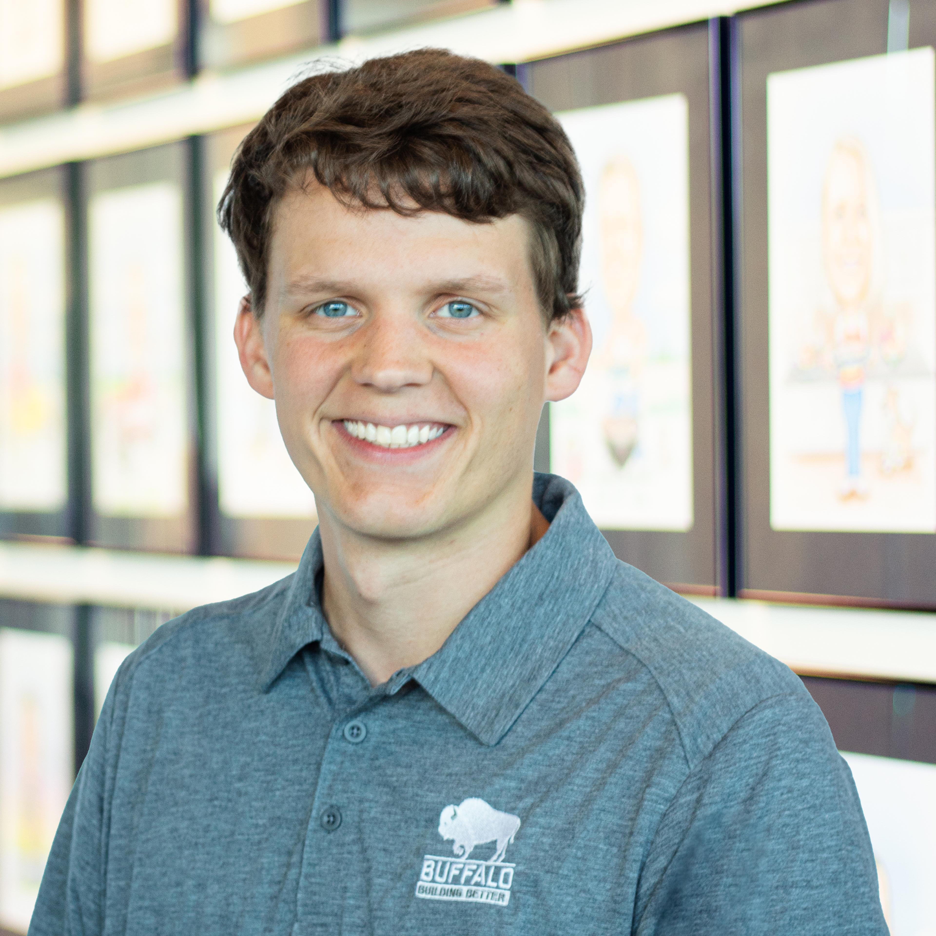 Young man with short brown hair and blue eyes in a gray Buffalo polo, smiling in front of framed illustrations.