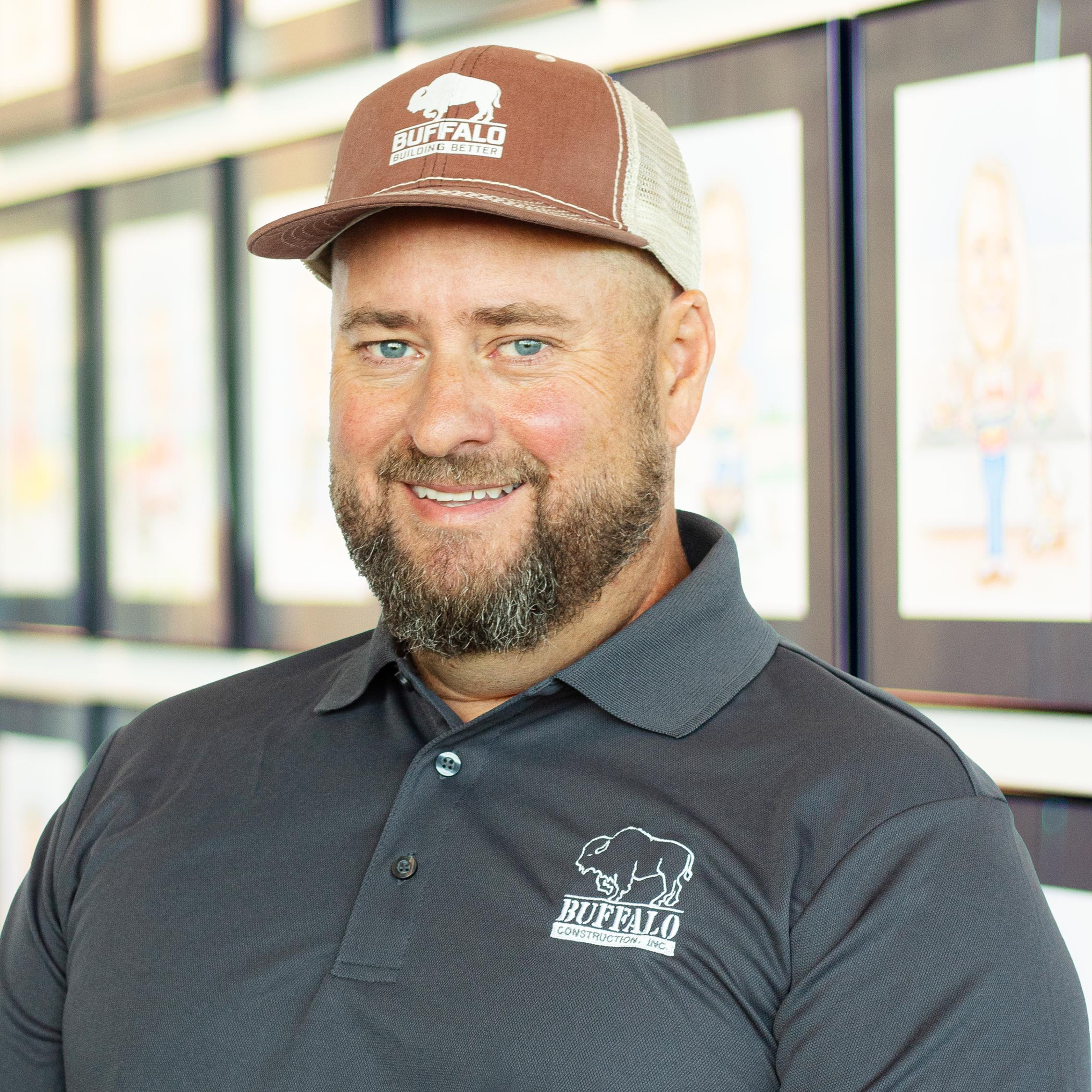 Clay Wallace wearing a buffalo branded polo and hat, standing in front of a wall displaying framed caricatures.