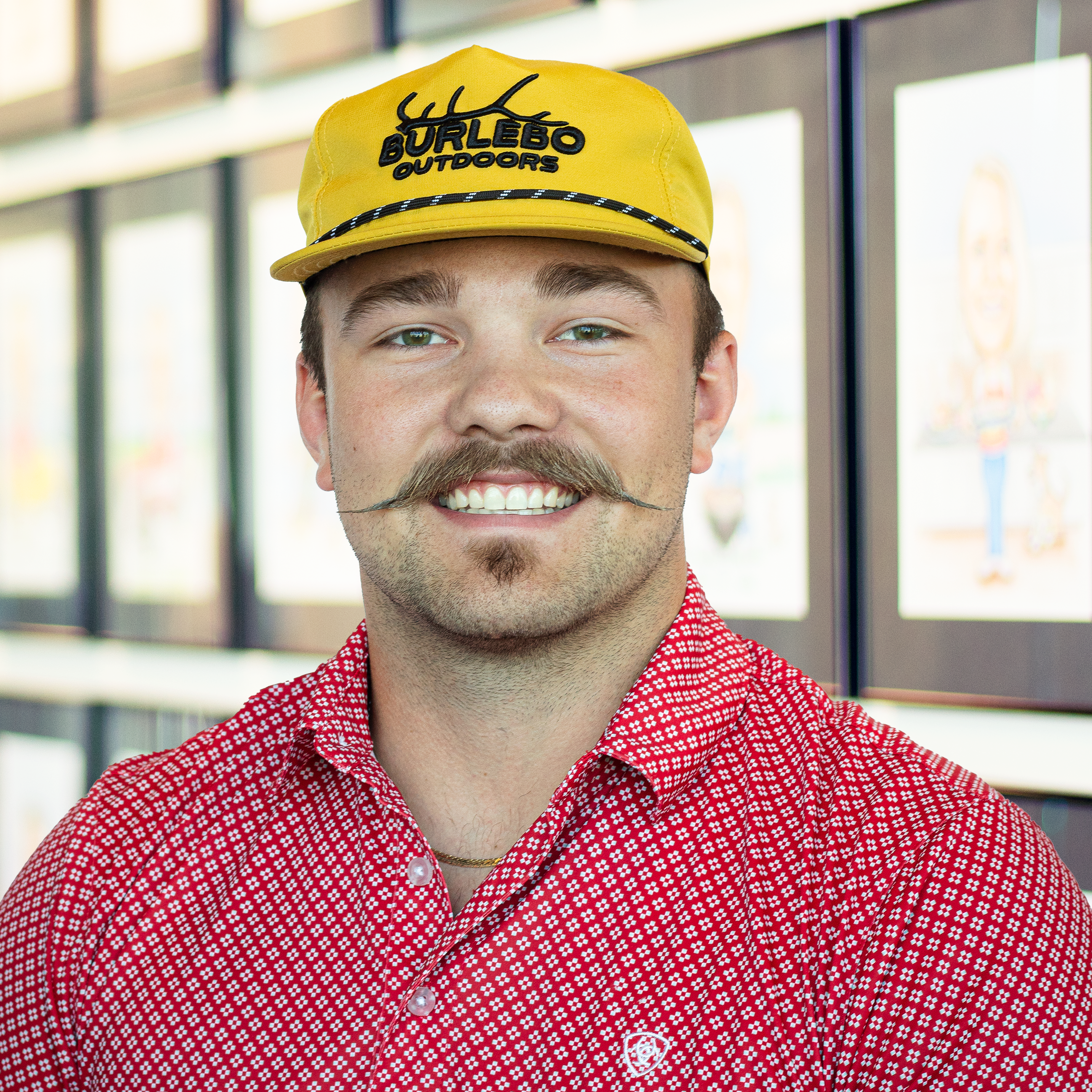 Blake Daher wearing a fun hat and red patterned polo, standing in front of a wall displaying framed caricatures.