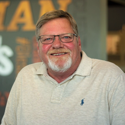 Chris Rogers wearing a light beige polo and glasses, standing in front of a company-branded wall.