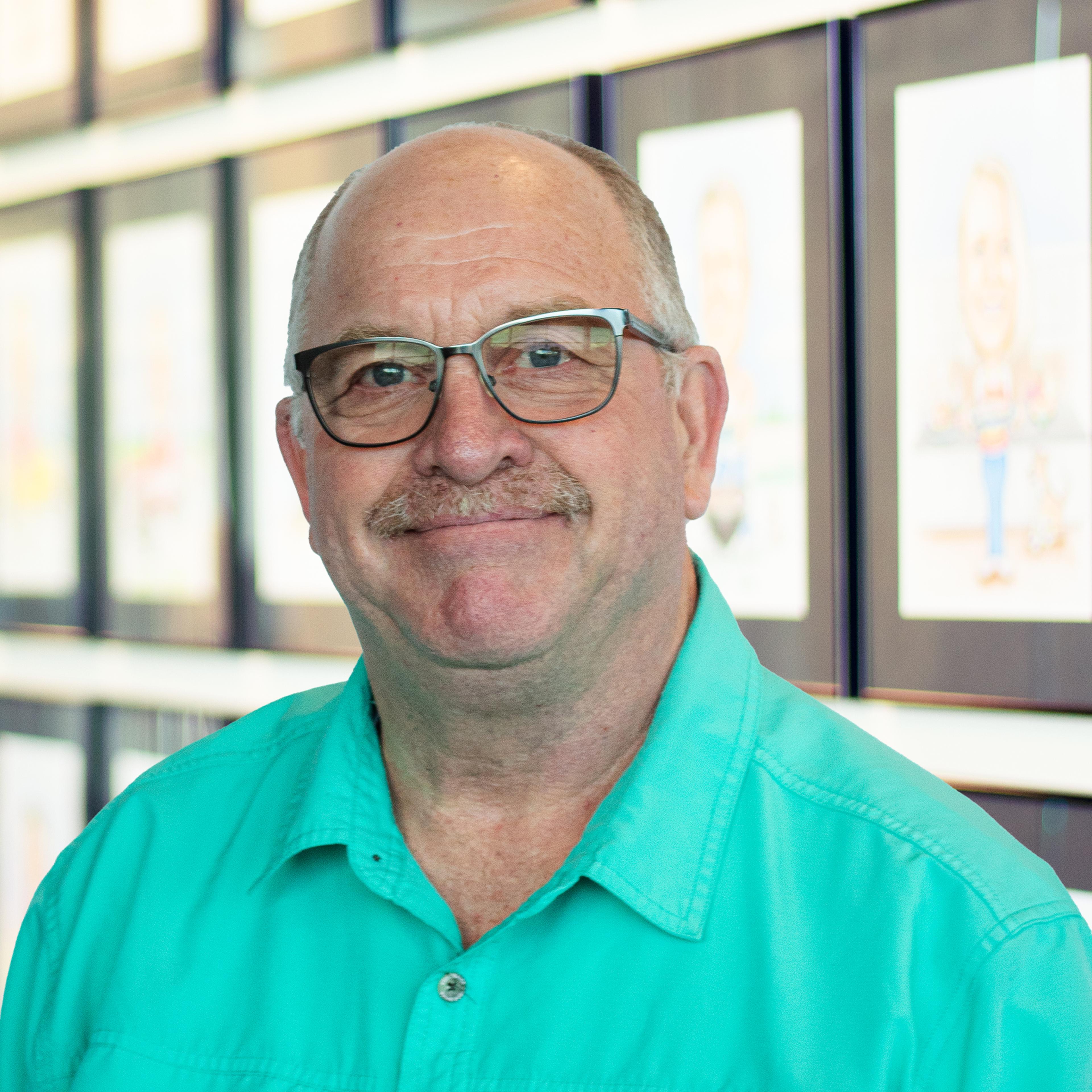 Tim Pelham, wearing a bright teal button-down shirt, standing in front of a wall displaying framed caricatures.