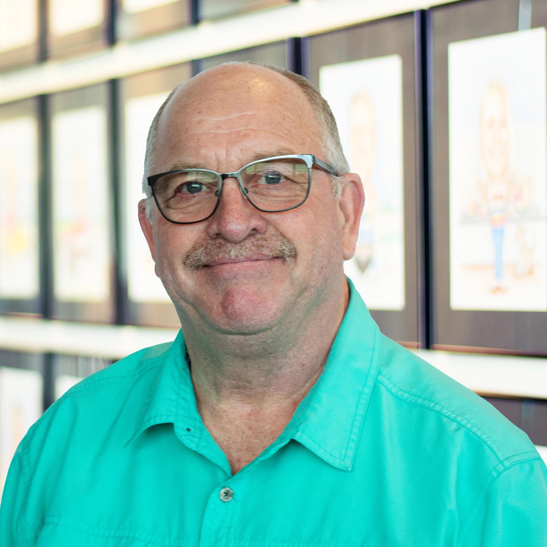 Tim Pelham, wearing a bright teal button-down shirt, standing in front of a wall displaying framed caricatures.