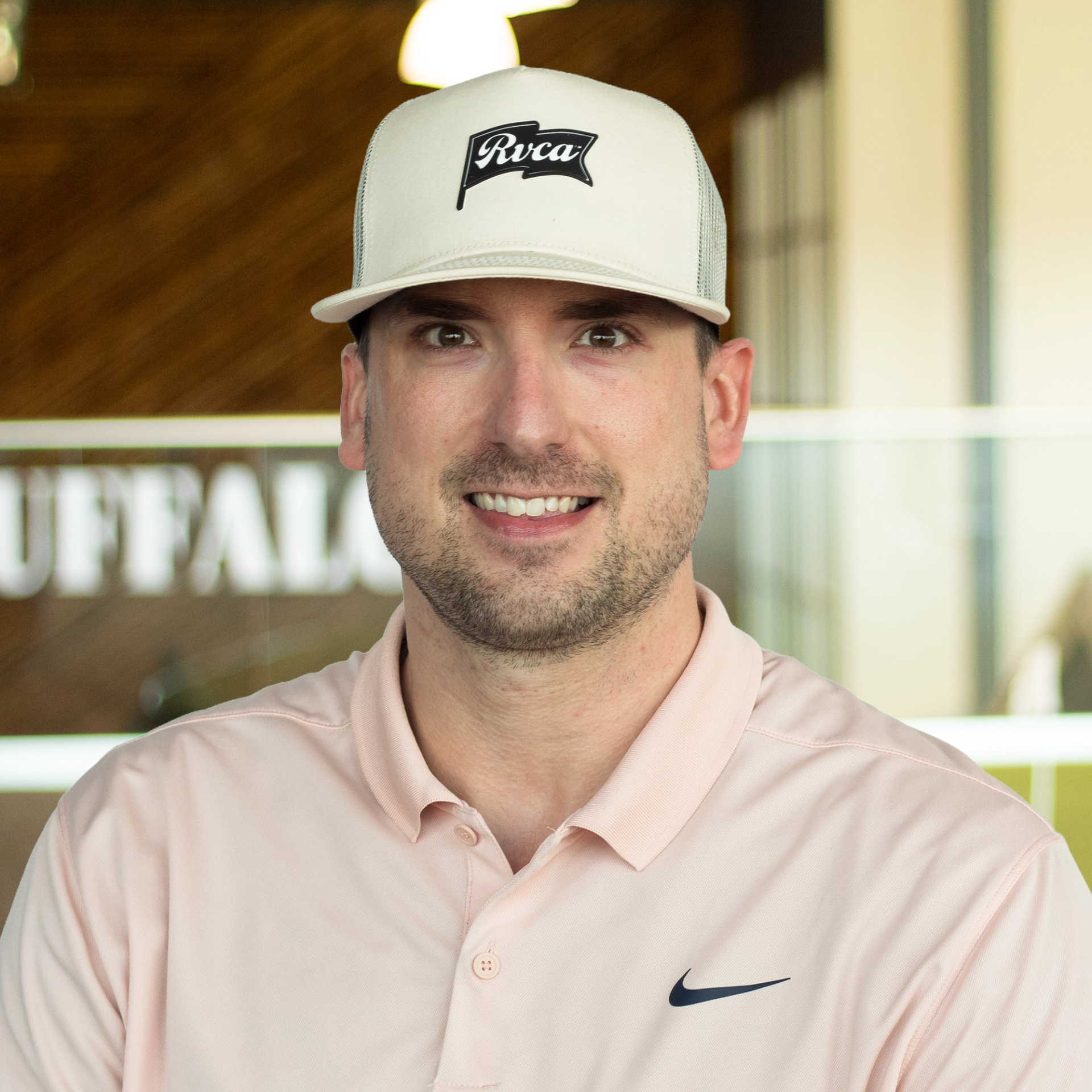 Collin Spink smiling, wearing a light pink polo and white hat, standing in front of a company-branded wall.