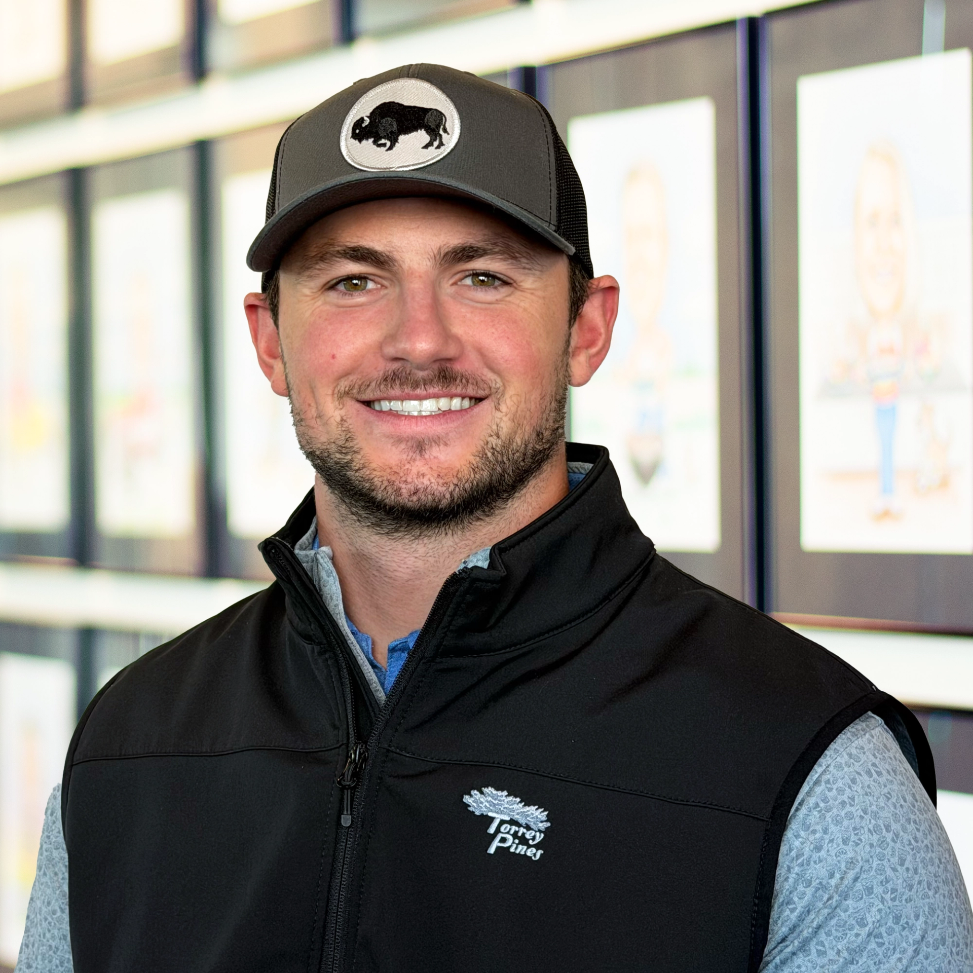 John Yeager, wearing a gray Buffalo hat and black T-shirt, standing in front of a wall displaying framed caricatures.