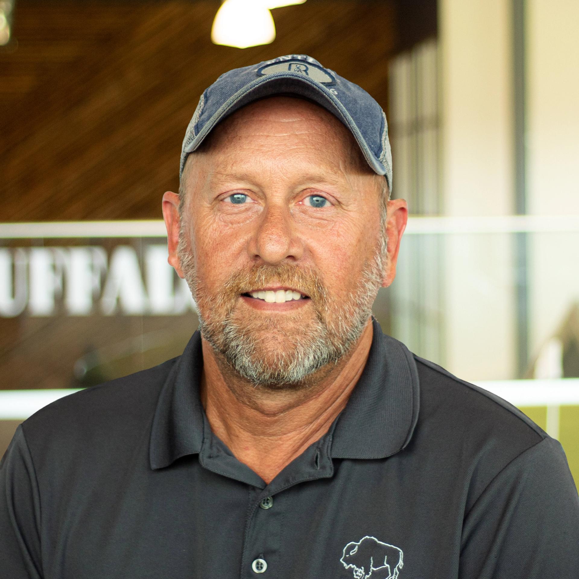 Brian Blank giving a hint of a smile, while wearing a gray buffalo branded polo standing in buffalo's office space.