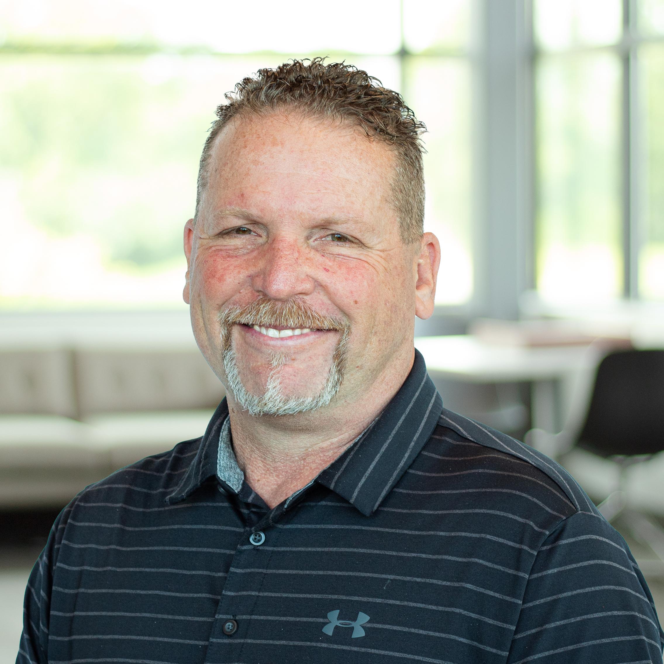 Man with short curly hair and a goatee in a black striped polo, smiling inside a bright, modern office.