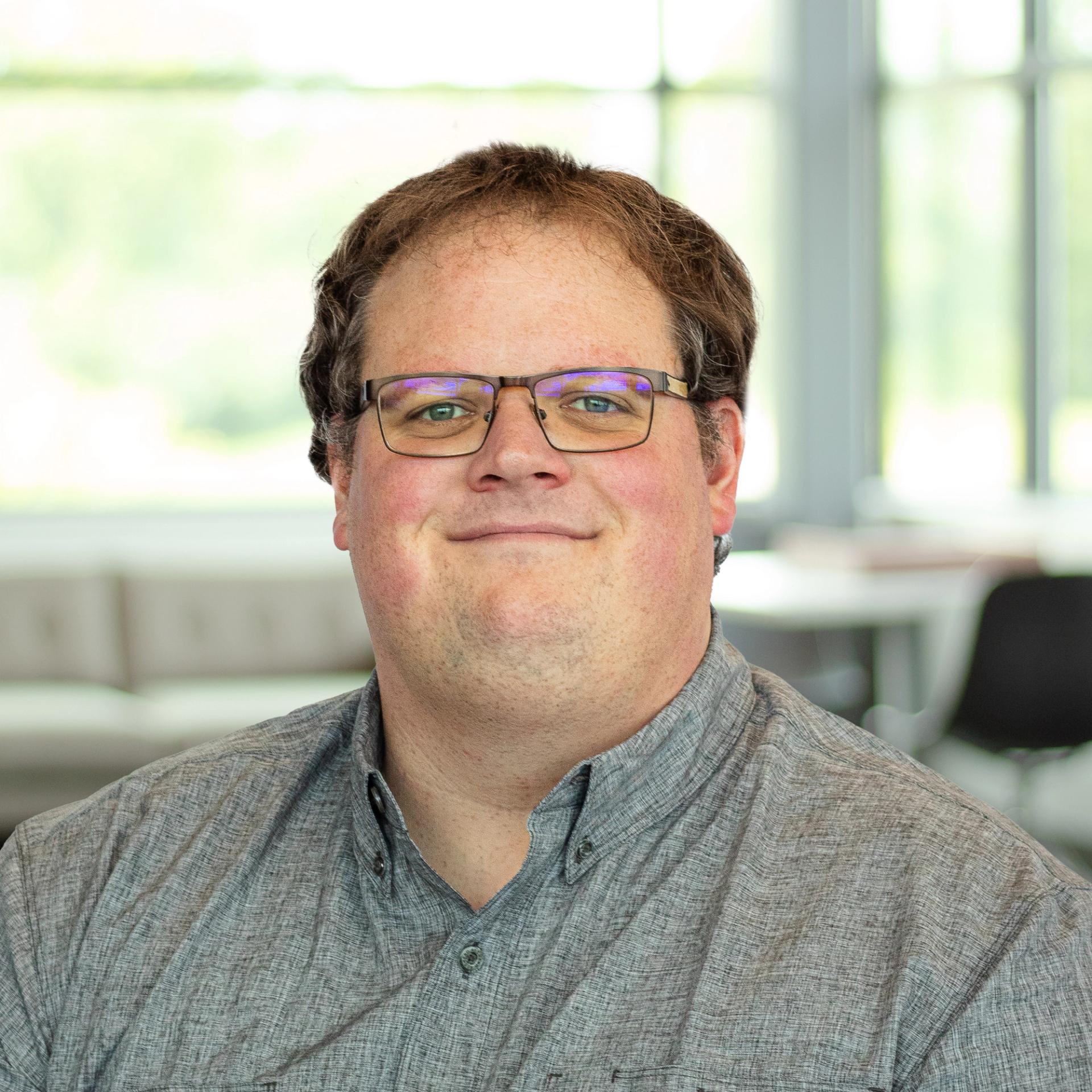 Joey Stephans wearing a gray shirt, smiling softly in a bright, modern office.