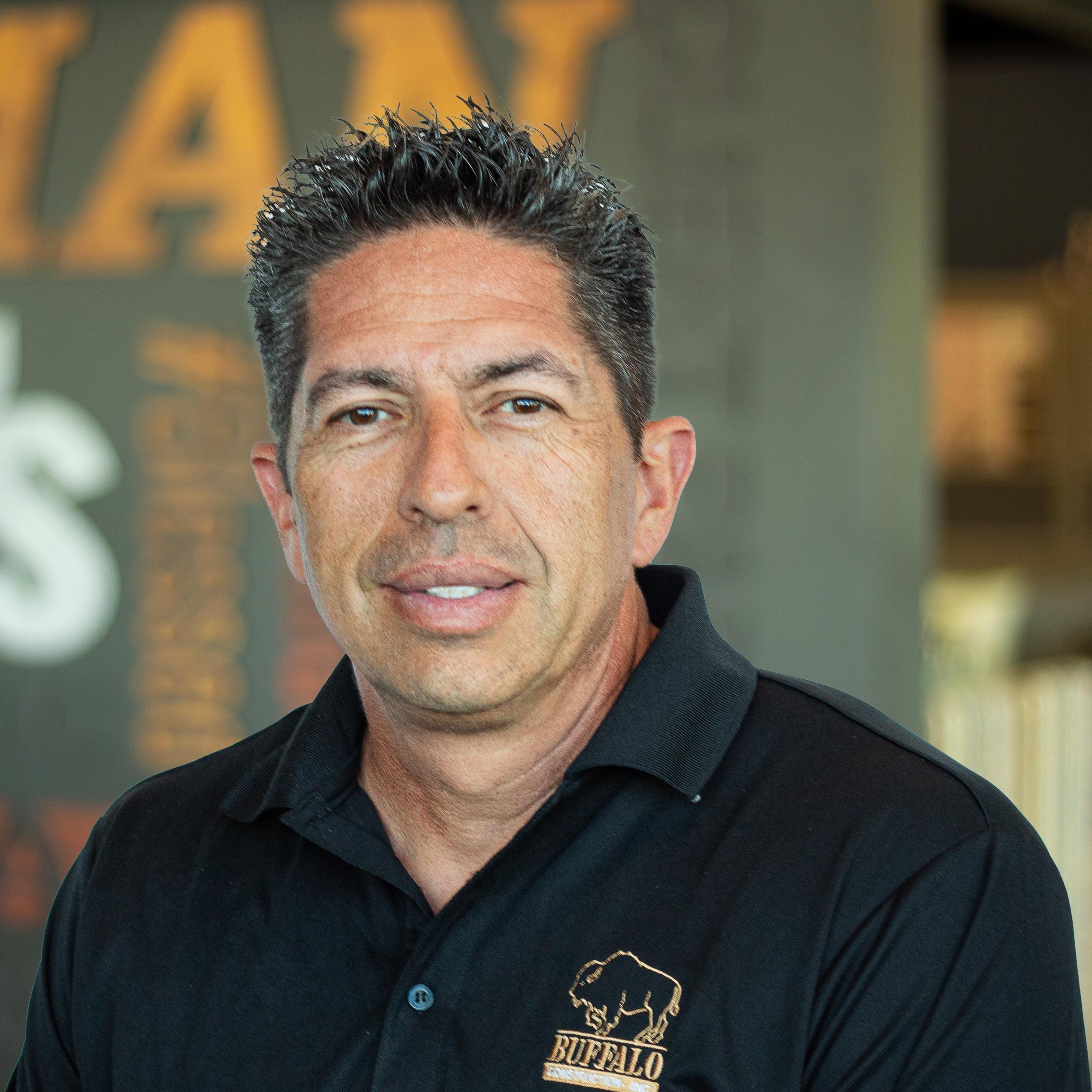 Man with spiked hair and a serious expression, wearing a black Buffalo polo, posing before a branded company wall.