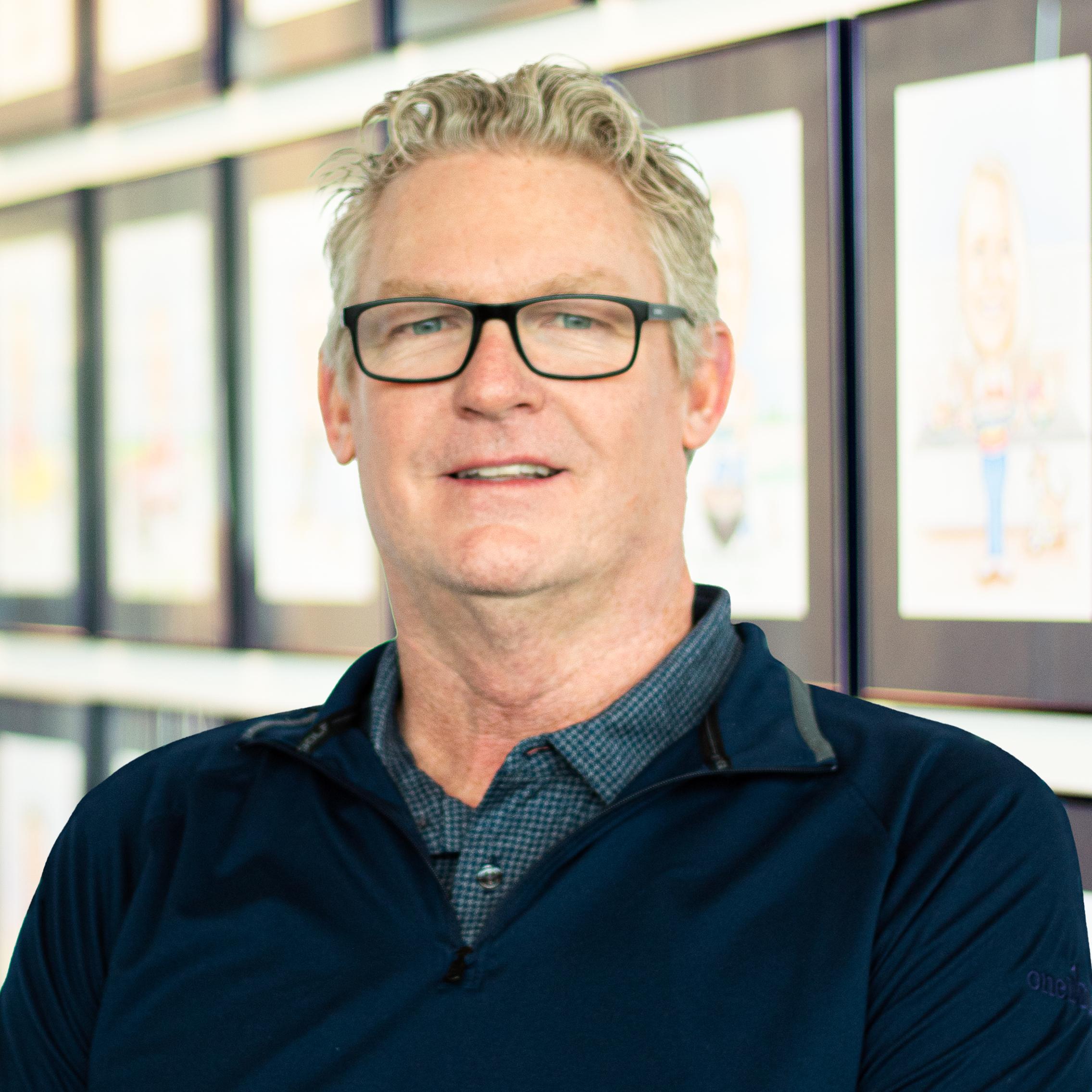 Andy Gregor wearing a smile, standing in front of a wall displaying framed caricatures.