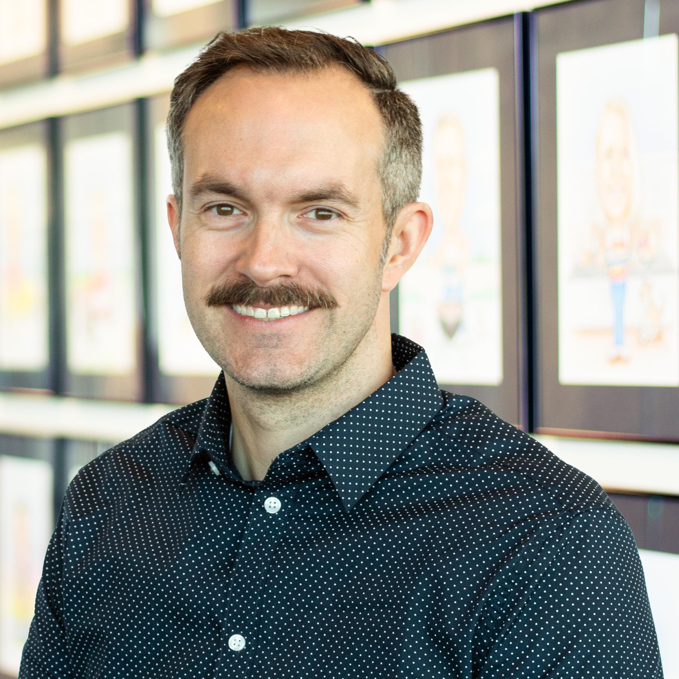 Matt Dudley wearing a large mustache and a smile while standing in front of a wall displaying framed caricatures.