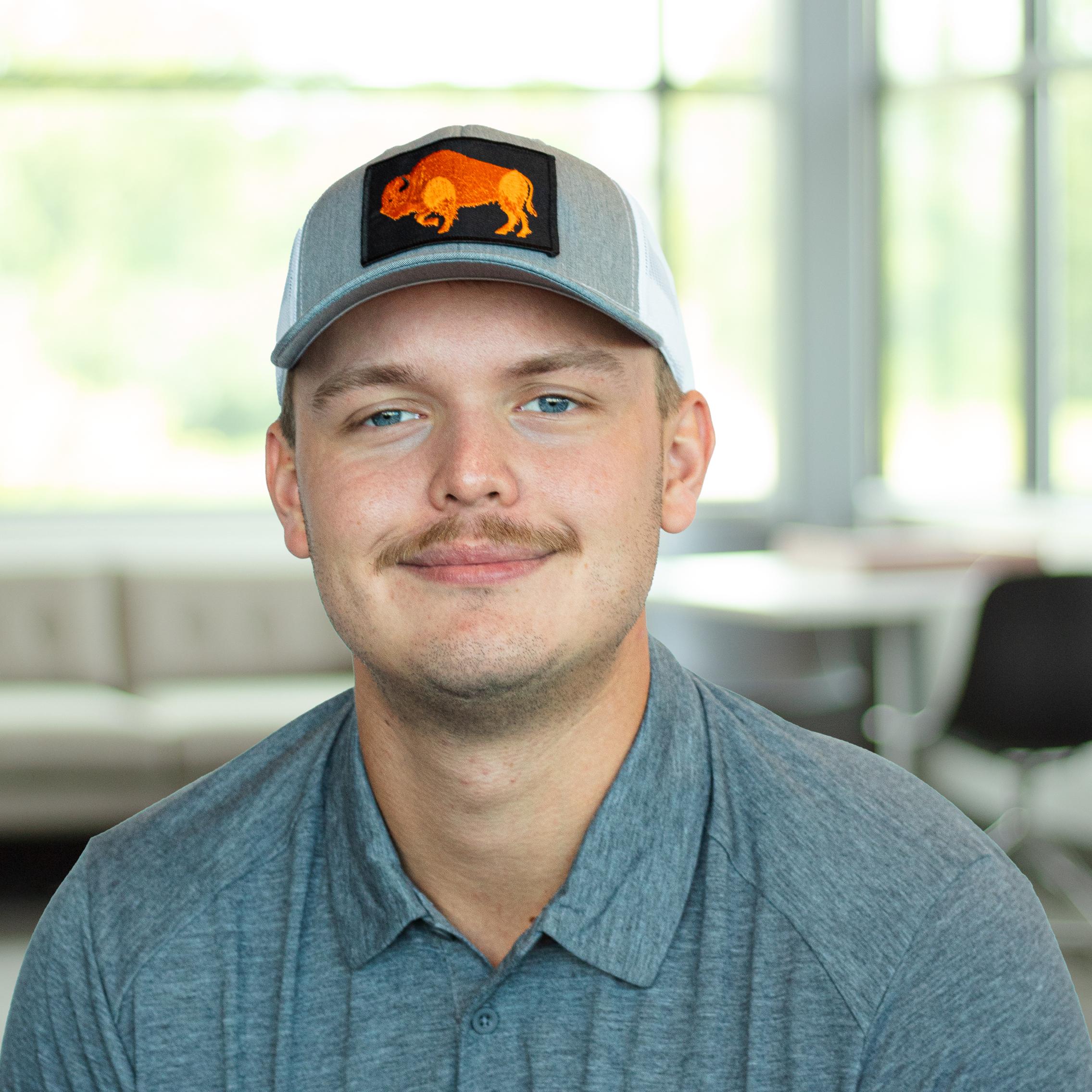 Sam Arthur, wearing a grey athletic polo and a grey mesh cap with an orange buffalo patch, smiling in a bright, modern office.