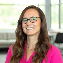 Kathryn Bell wearing a bright pink blouse and glasses, smiling in a bright, modern office.