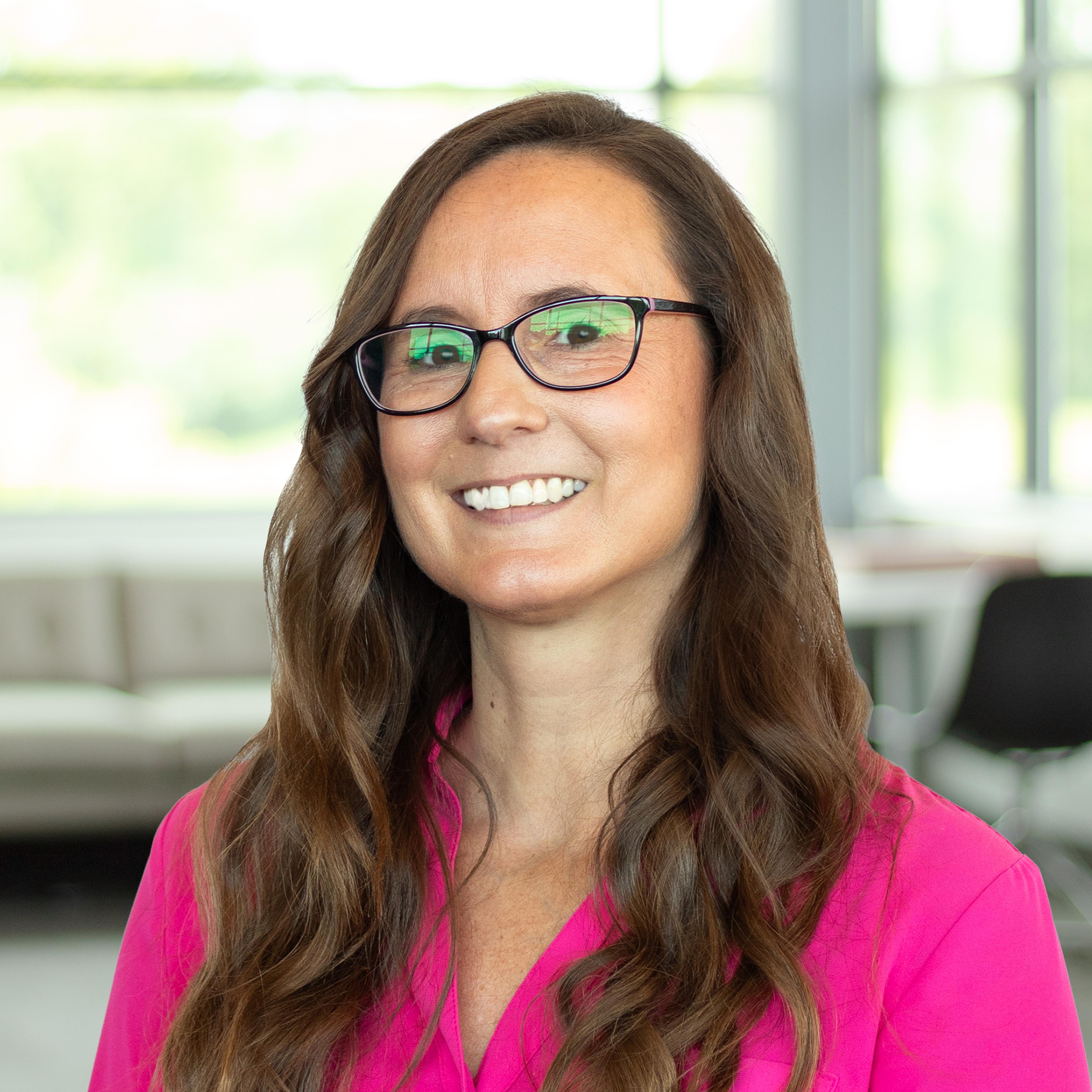 Kathryn Bell wearing a bright pink blouse and glasses, smiling in a bright, modern office.