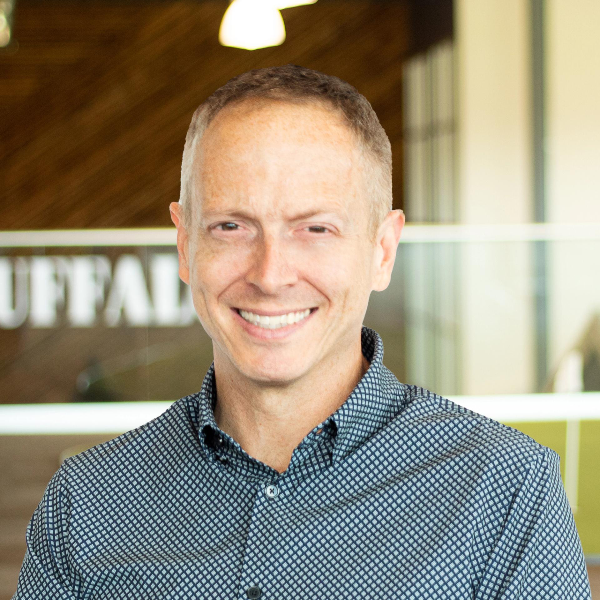 Brett Norton smiling, wearing a nice button down, standing in buffalo's office space.
