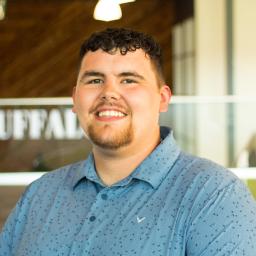 Zach Tong smiles while wearing a light blue patterned button-down shirt, standing in the Buffalo Construction office with a wood accent wall in the background.