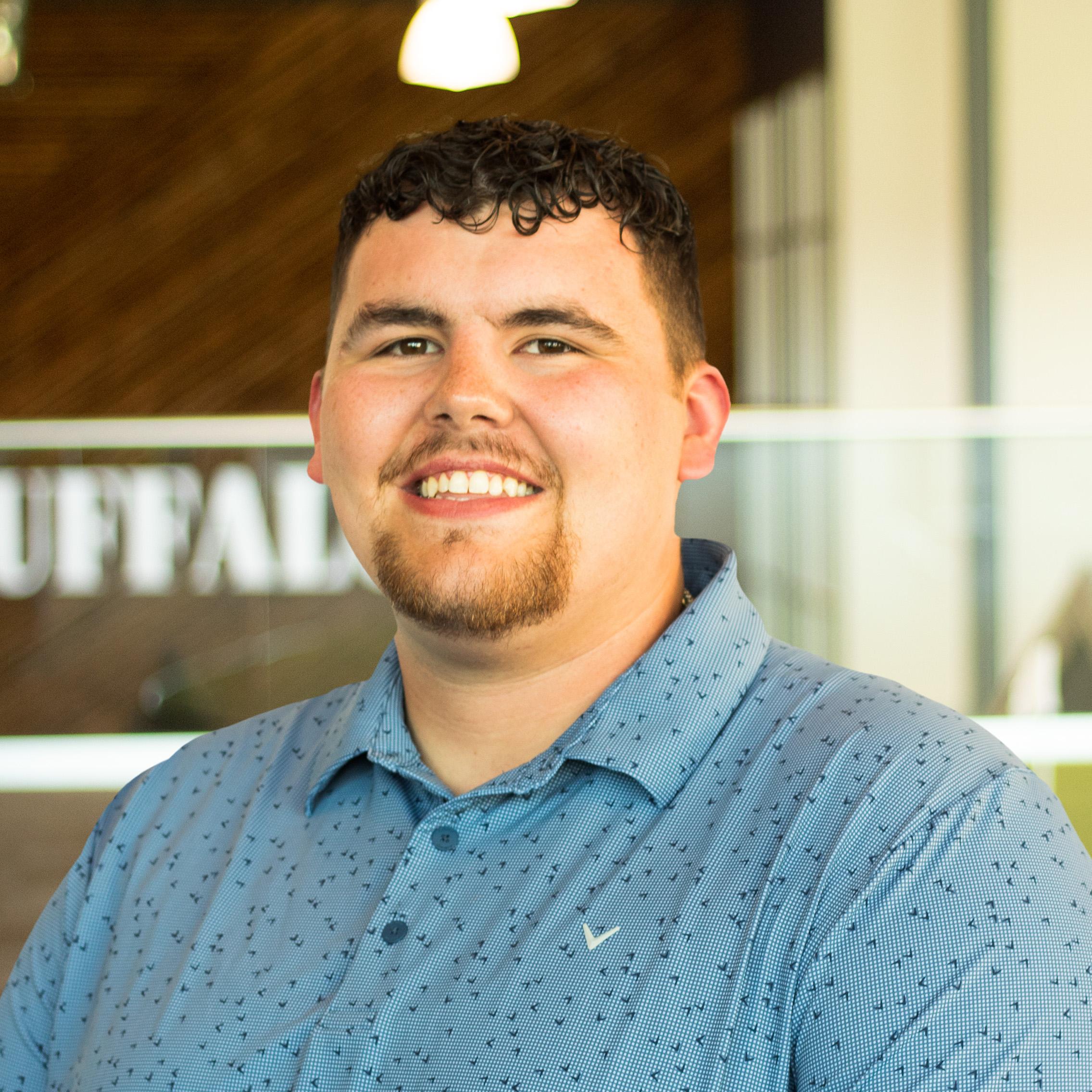 Zach Tong smiles while wearing a light blue patterned button-down shirt, standing in the Buffalo Construction office with a wood accent wall in the background.