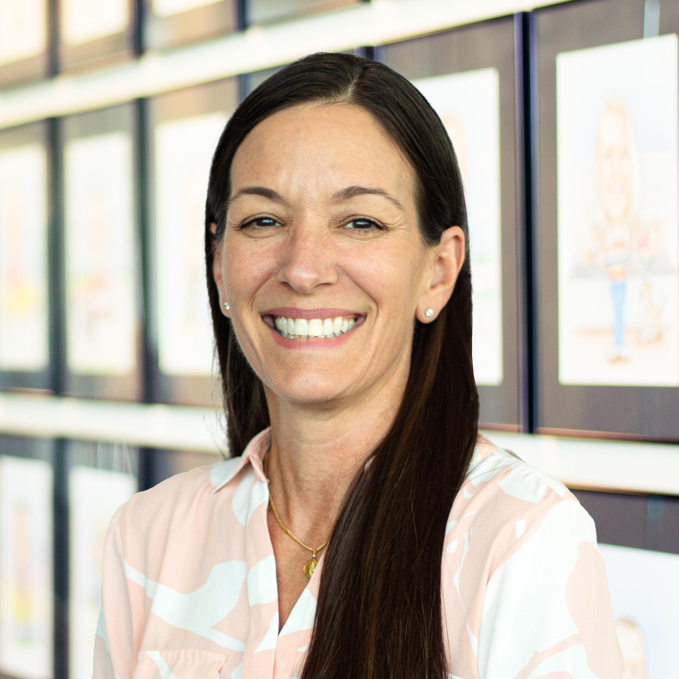 Christine Rey wearing a bright smile and standing in front of a wall displaying framed caricatures.