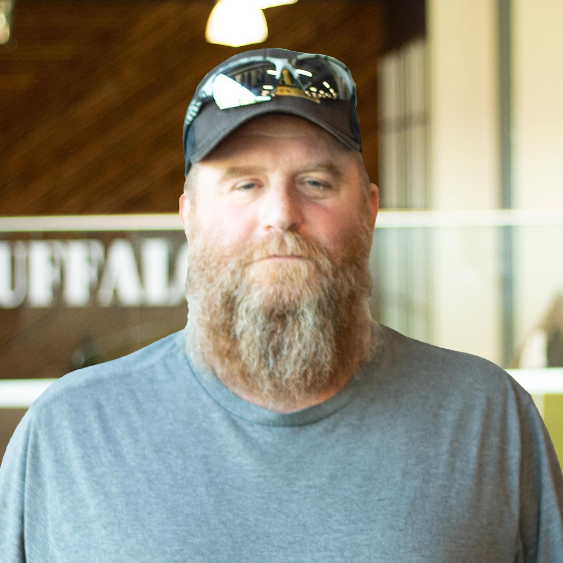 Greg Riggs with a serious face, wearing a gray t-shirt and standing in buffalo's office space.