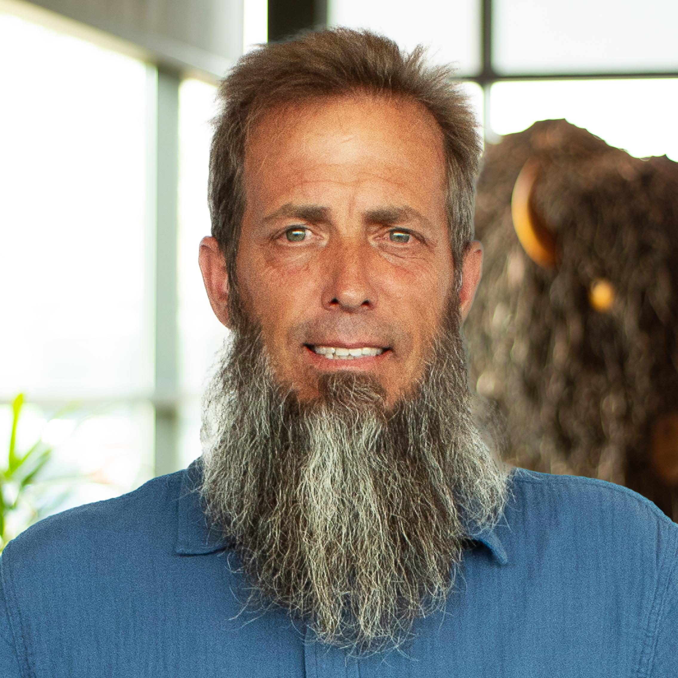 Steve Oyler with a long beard and a nice smile standing in front of a company-branded wall.