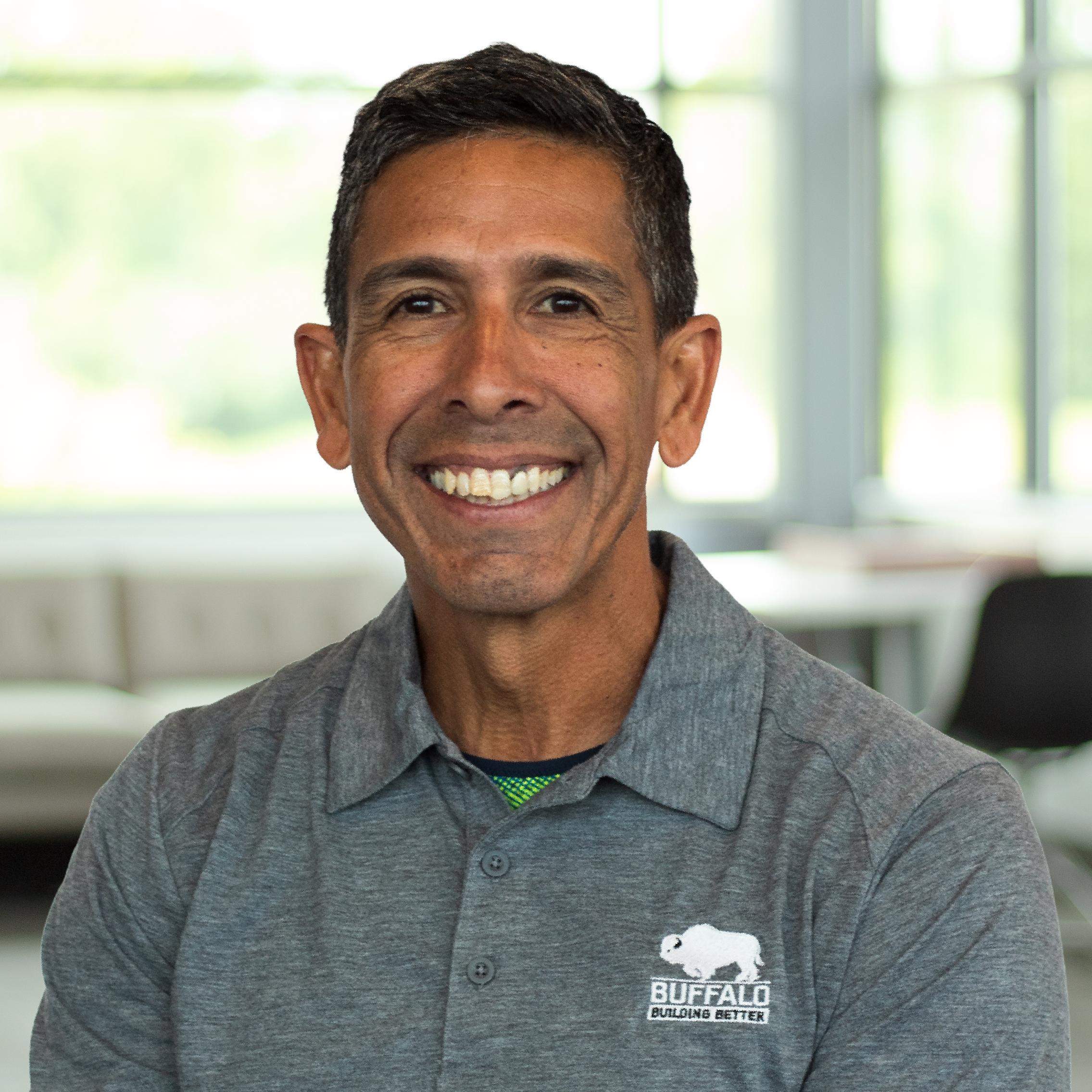 Enrique Mercado smiling, wearing a gray Buffalo polo, standing in a bright office space.