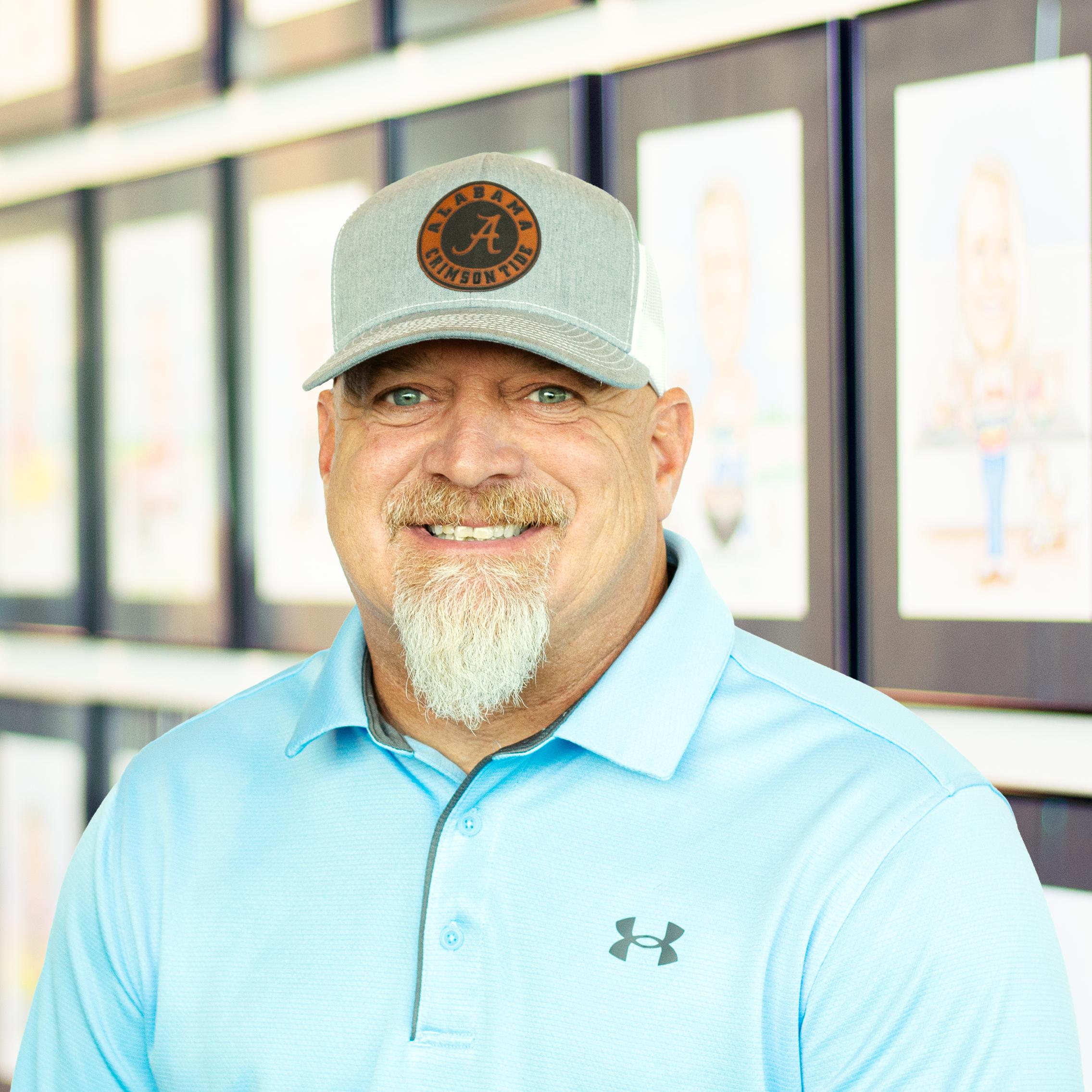 Jason Dillard with bright blue eyes and a smile, standing in front of a wall displaying framed caricatures.