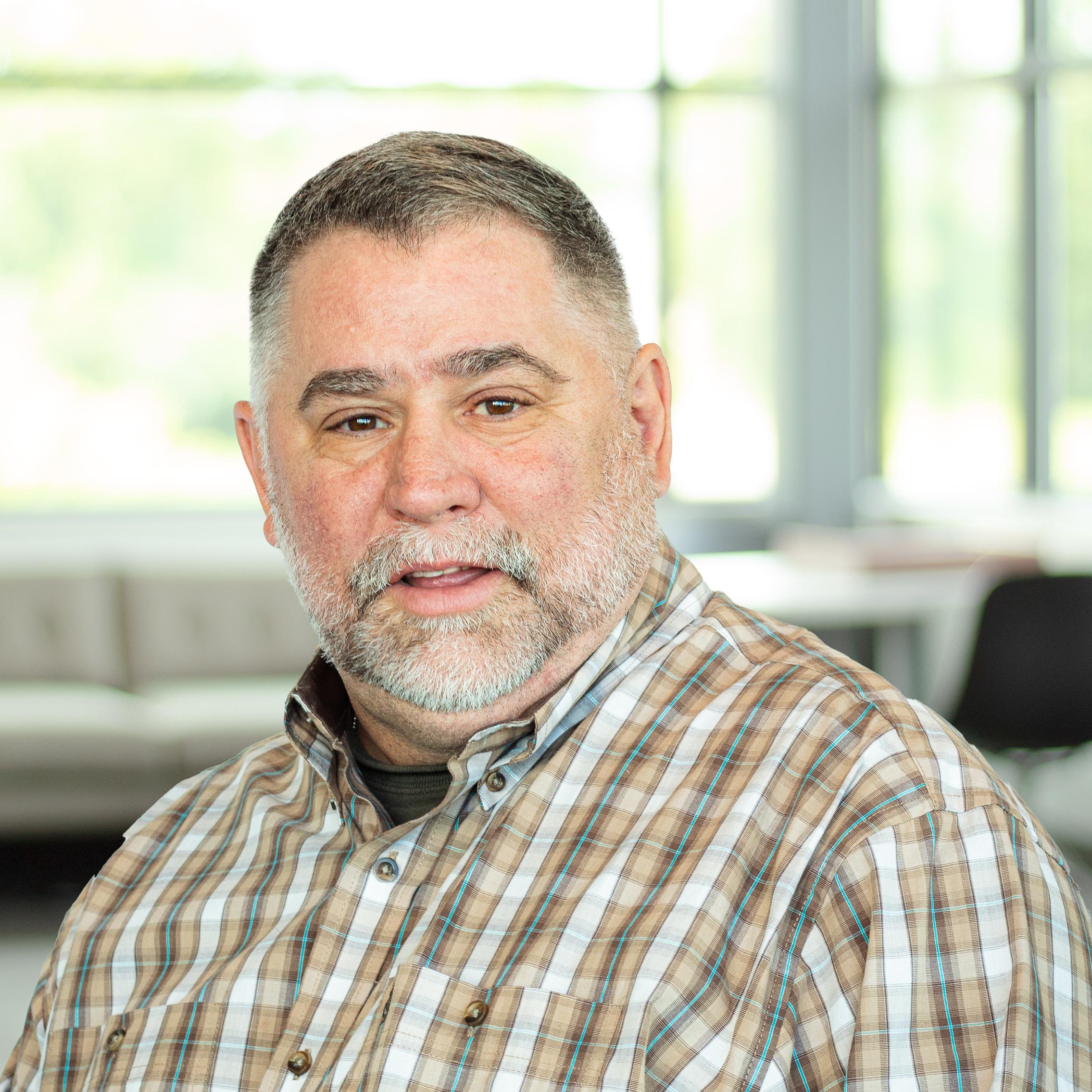 Bearded man in a plaid shirt with a neutral expression, standing inside a well-lit modern office lounge.