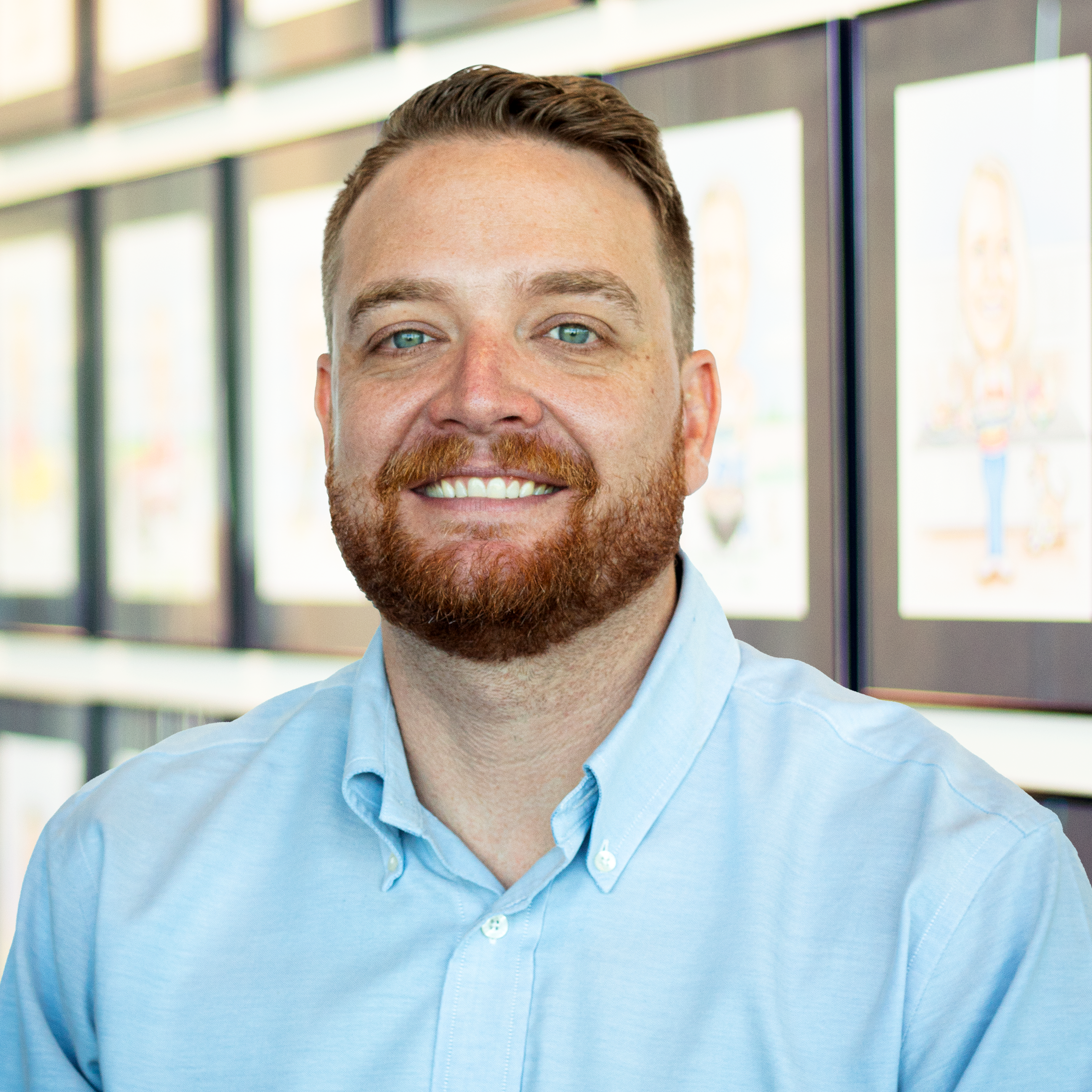 Colby Corbell with bright blue eyes, smiling and standing in front of a wall displaying framed caricatures.