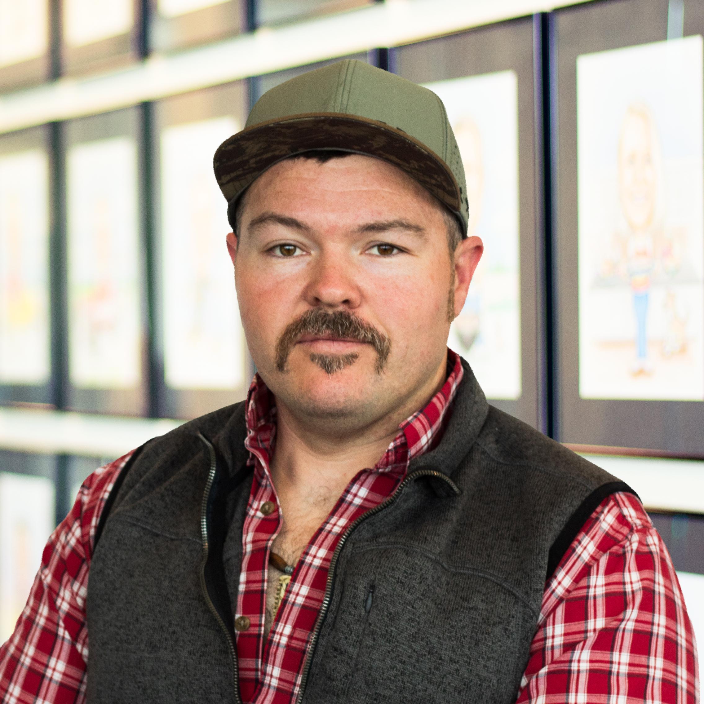 Michael Simpson, wearing a red plaid shirt and dark vest, standing in front of a wall displaying framed caricatures.