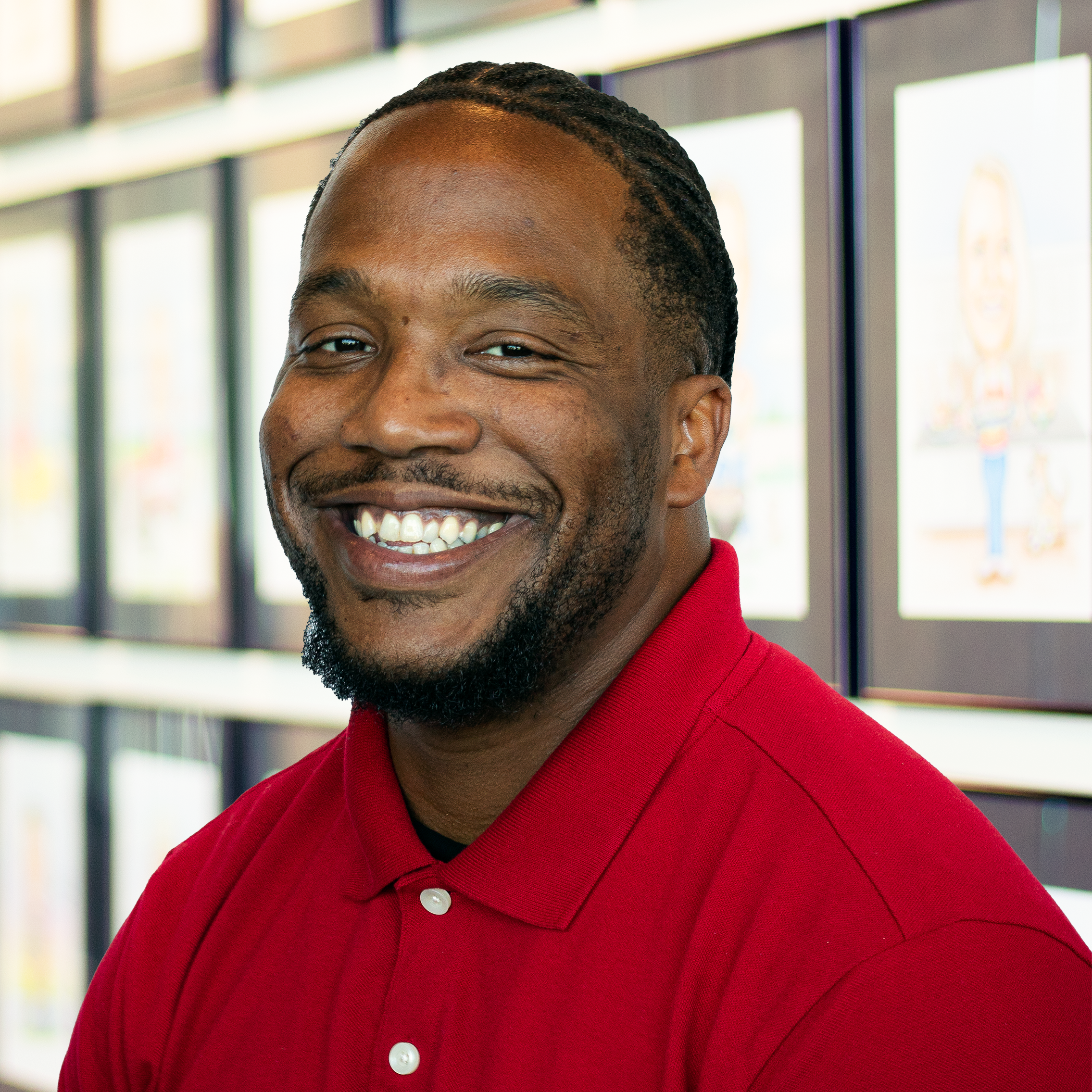 Aaron Fowler wearing a red polo, smiling and standing in front of a wall displaying framed caricatures.