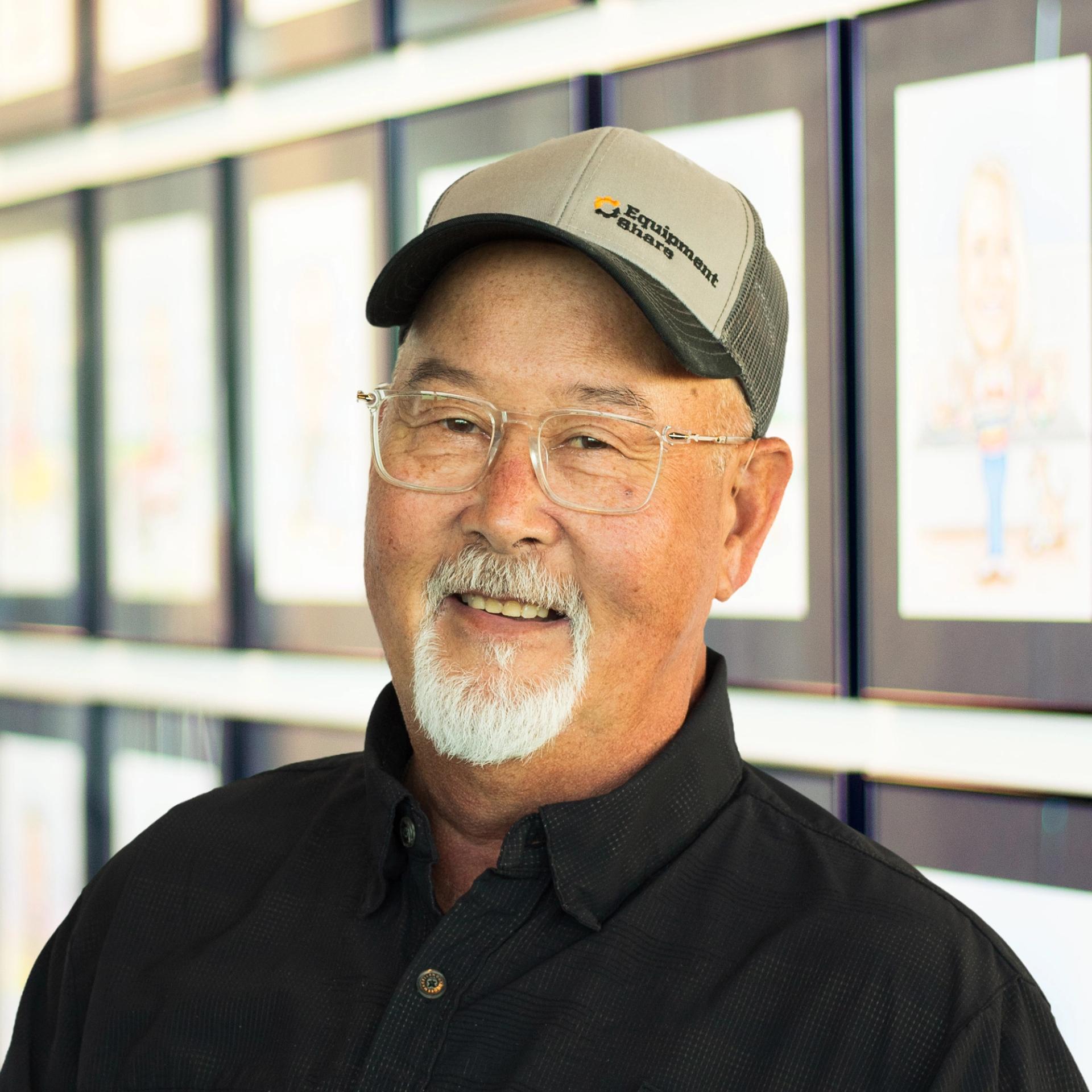 David Kilgore smiles while wearing a black button-down shirt, gray EquipmentShare cap, and glasses, standing in front of a wall displaying framed caricatures.