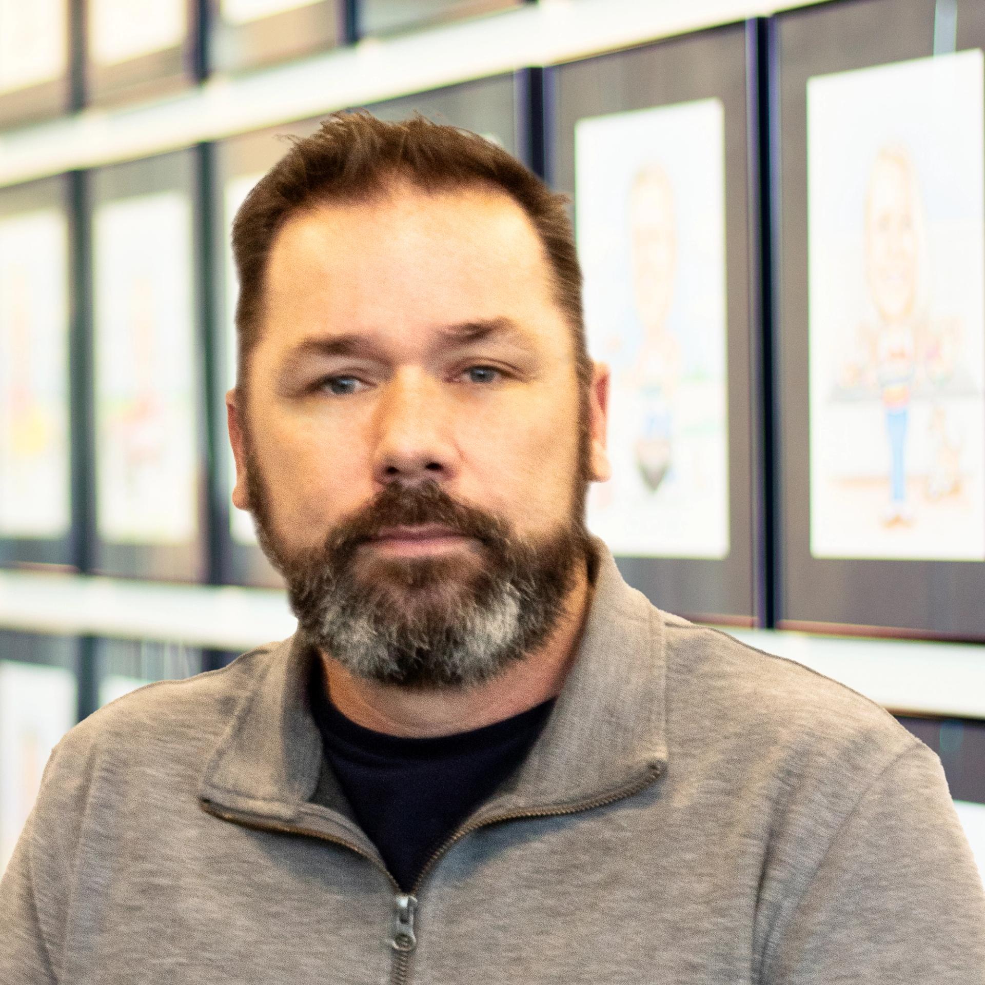 Bill McCrary wearing a great sweater and standing in front of a wall displaying framed caricatures.