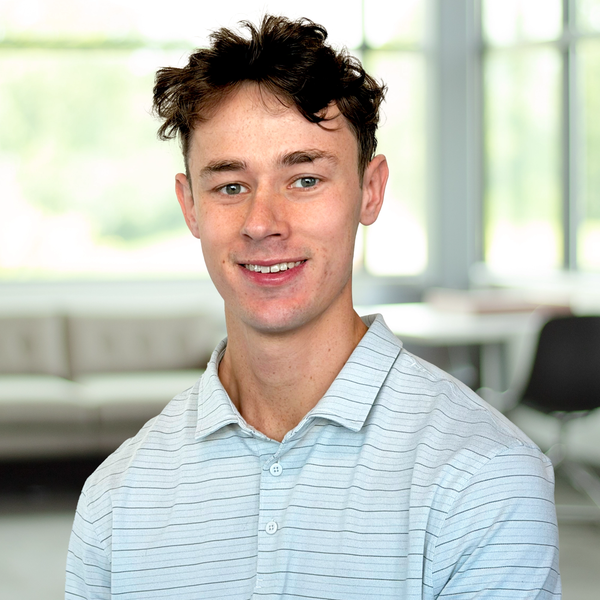 Matthew Schneider wearing a light blue striped polo, smiling in a bright, modern office.