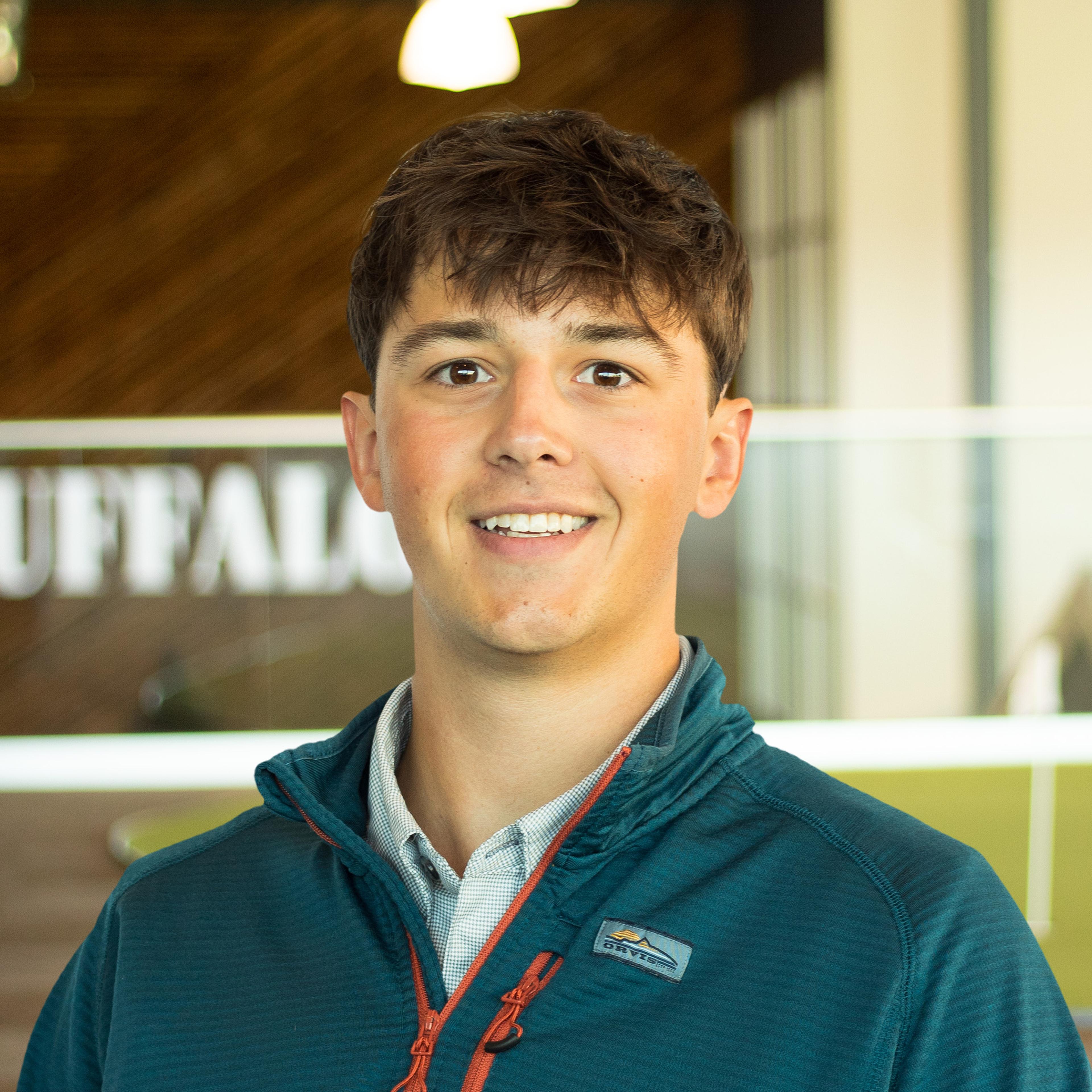 Carson Elmore smiles while wearing a teal quarter-zip over a button-down shirt, standing in the Buffalo Construction office with a wood accent wall in the background.