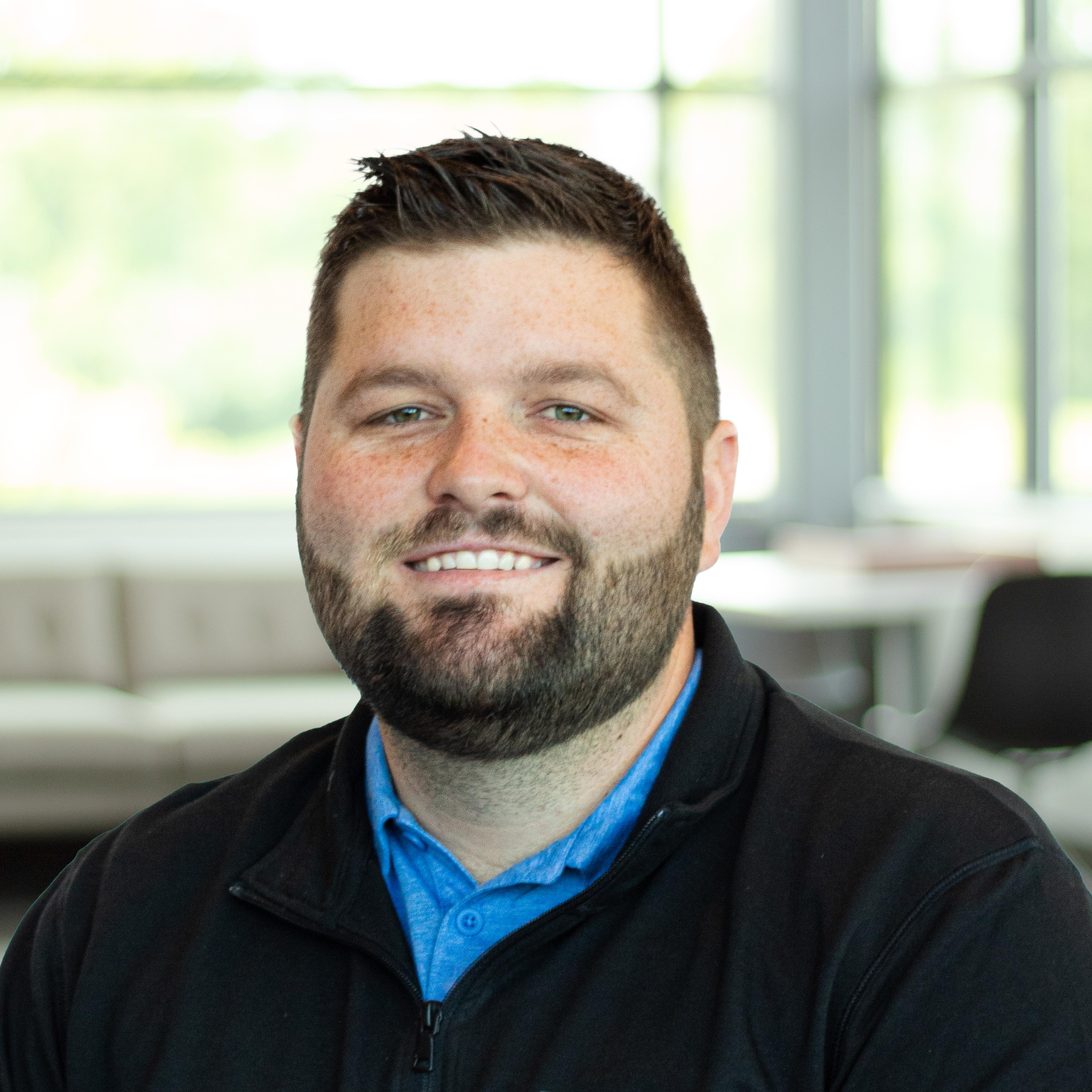 Blade Kearns wearing a black sweater and blue shirt, smiling in a bright, modern office.