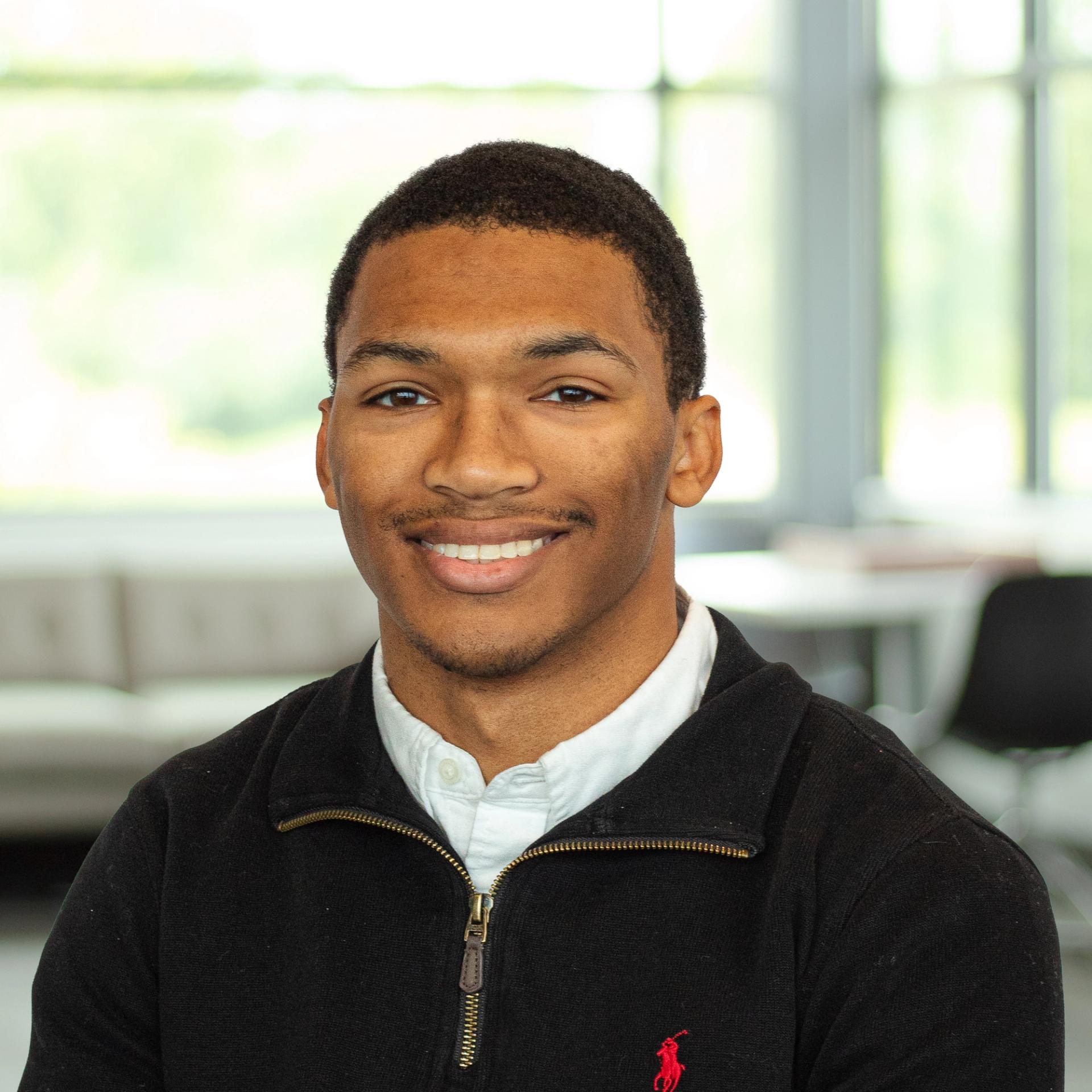 Caleb Griggs wearing a black sweater with a white shirt, smiling confidently in a bright, modern office.