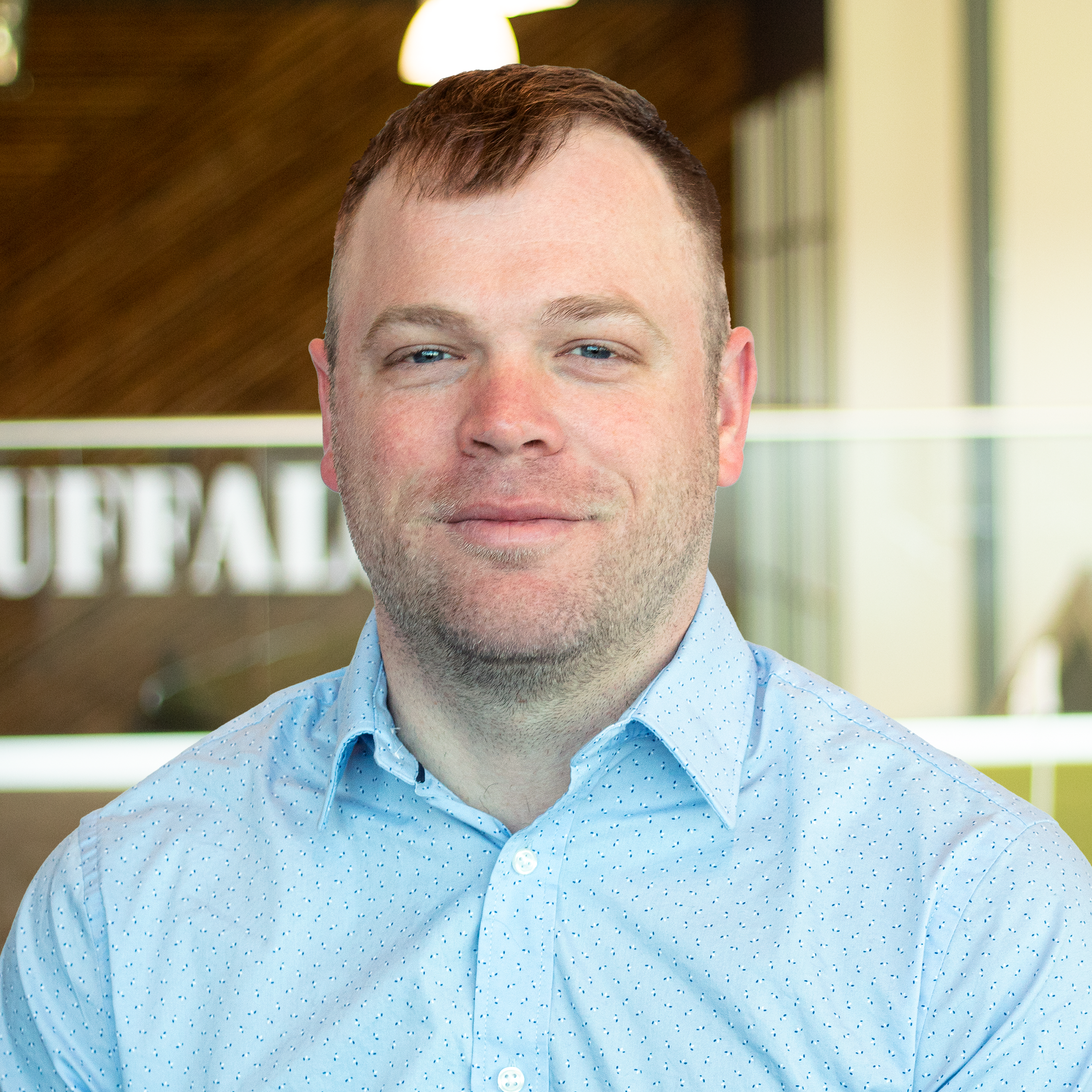 Blake Williams with a faint smile, wearing a light-blue button down, standing in buffalo's office space.