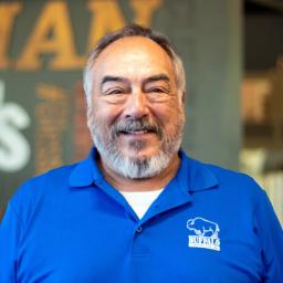 Enos Colvin wearing a bright blue buffalo branded polo, smiling and standing in front of a company-branded wall.